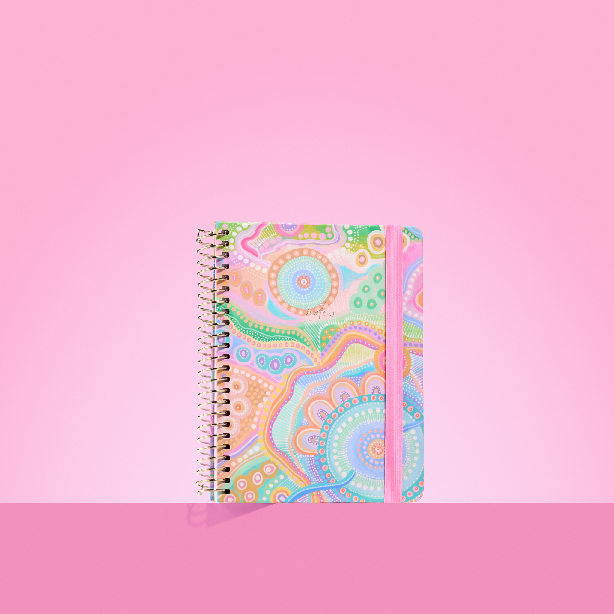 Pastel-toned 2026 notepad with Aboriginal-inspired circular patterns, gold spiral binding, and pink elastic closure on a pink background.