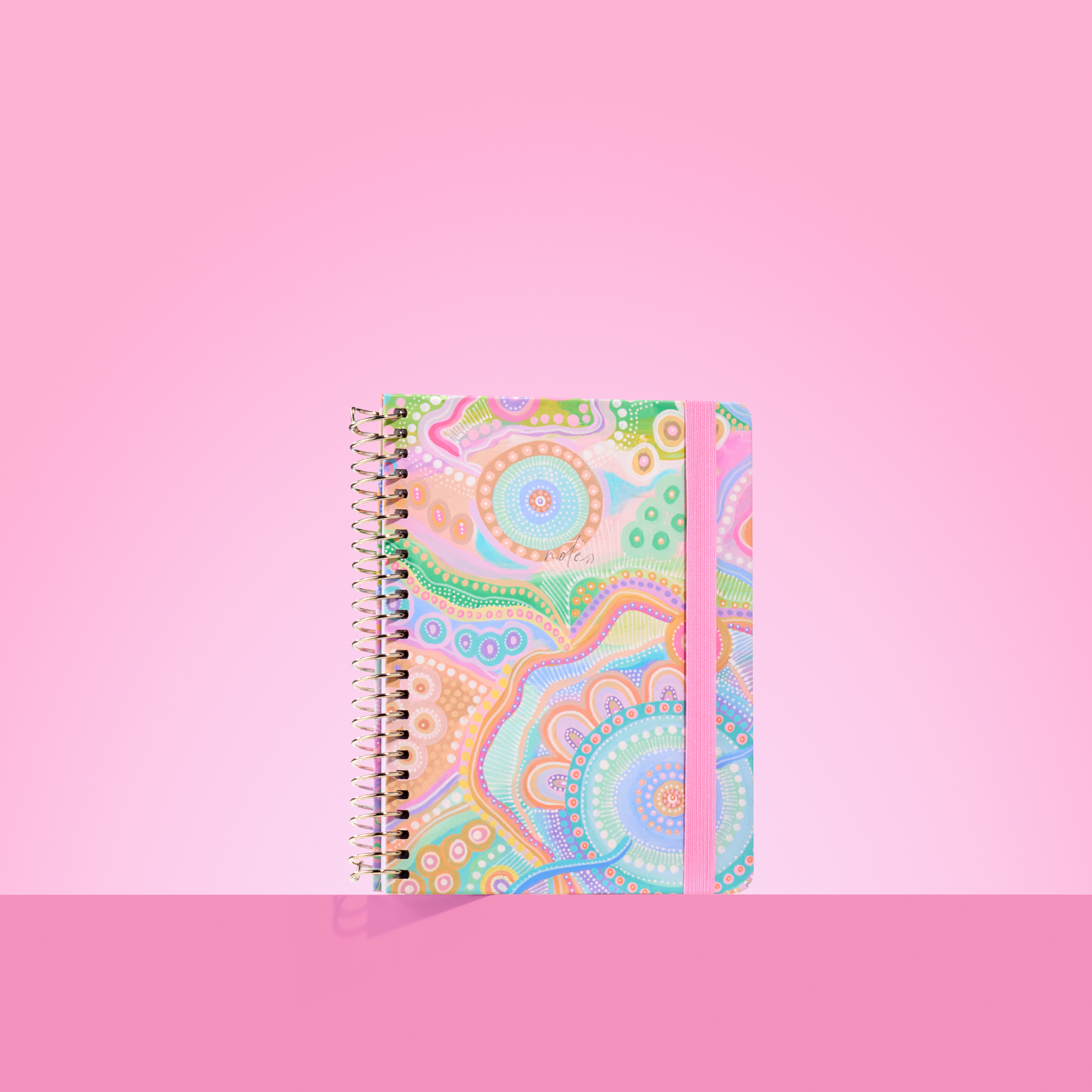 Pastel-toned 2026 notepad with Aboriginal-inspired circular patterns, gold spiral binding, and pink elastic closure on a pink background.