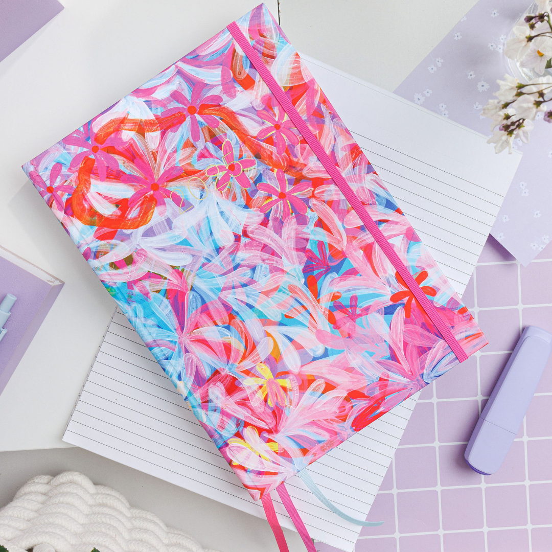 Floral 'Upstream' weekly planner with warm tones, resting flat on a lined notebook on a desk, showcasing its vibrant, nature-inspired artwork.