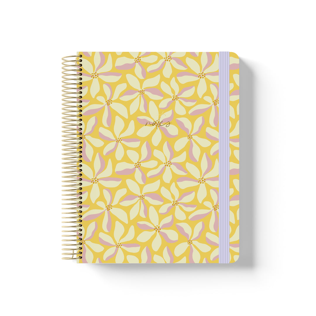 Yellow coiled notebook with gold spiral, floral petal pattern in pink and cream, lavender elastic band, and “notes” in gold script.