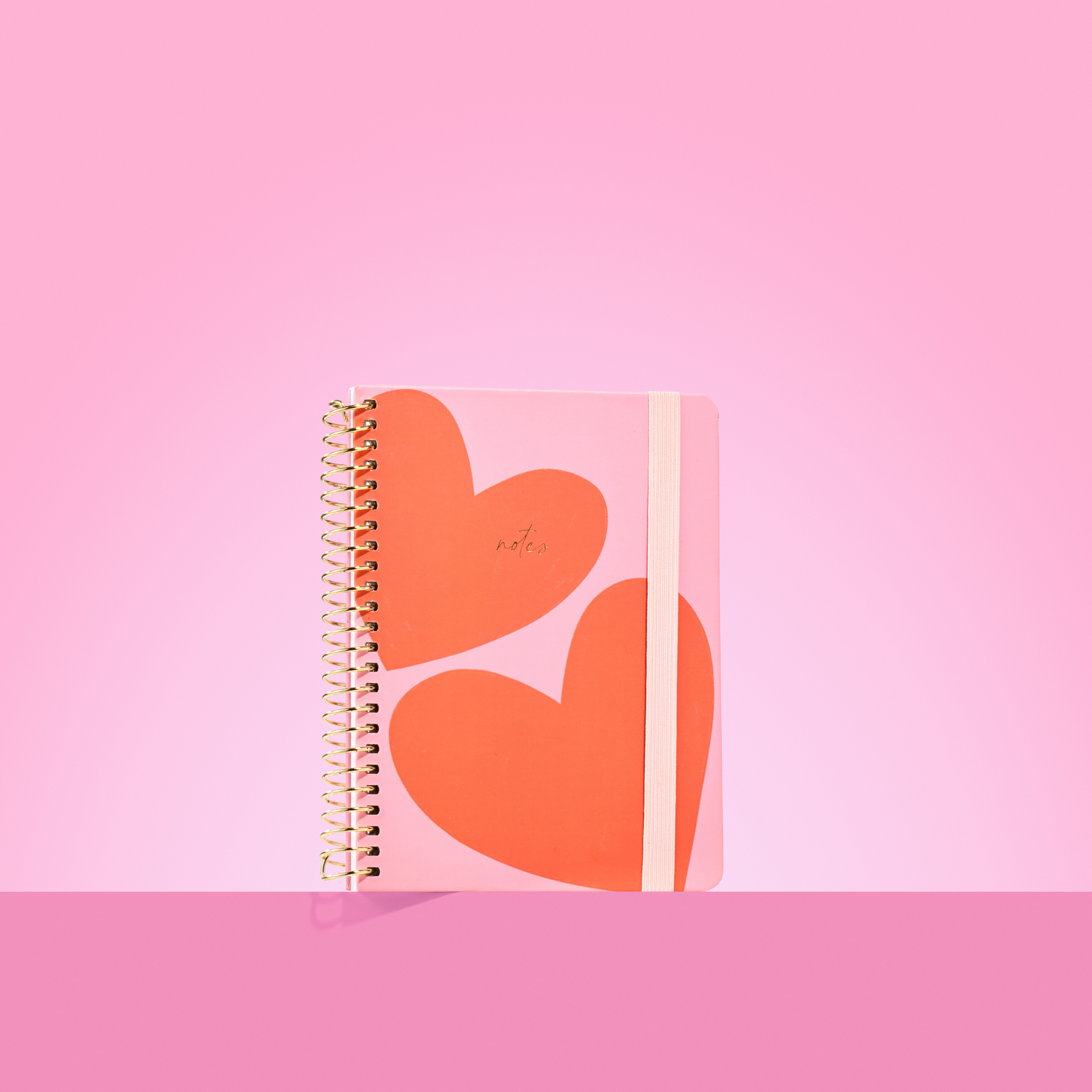 Pink 2026 notepad with two large red-orange hearts, gold spiral binding, light pink elastic closure, and "notes" embossed in gold.