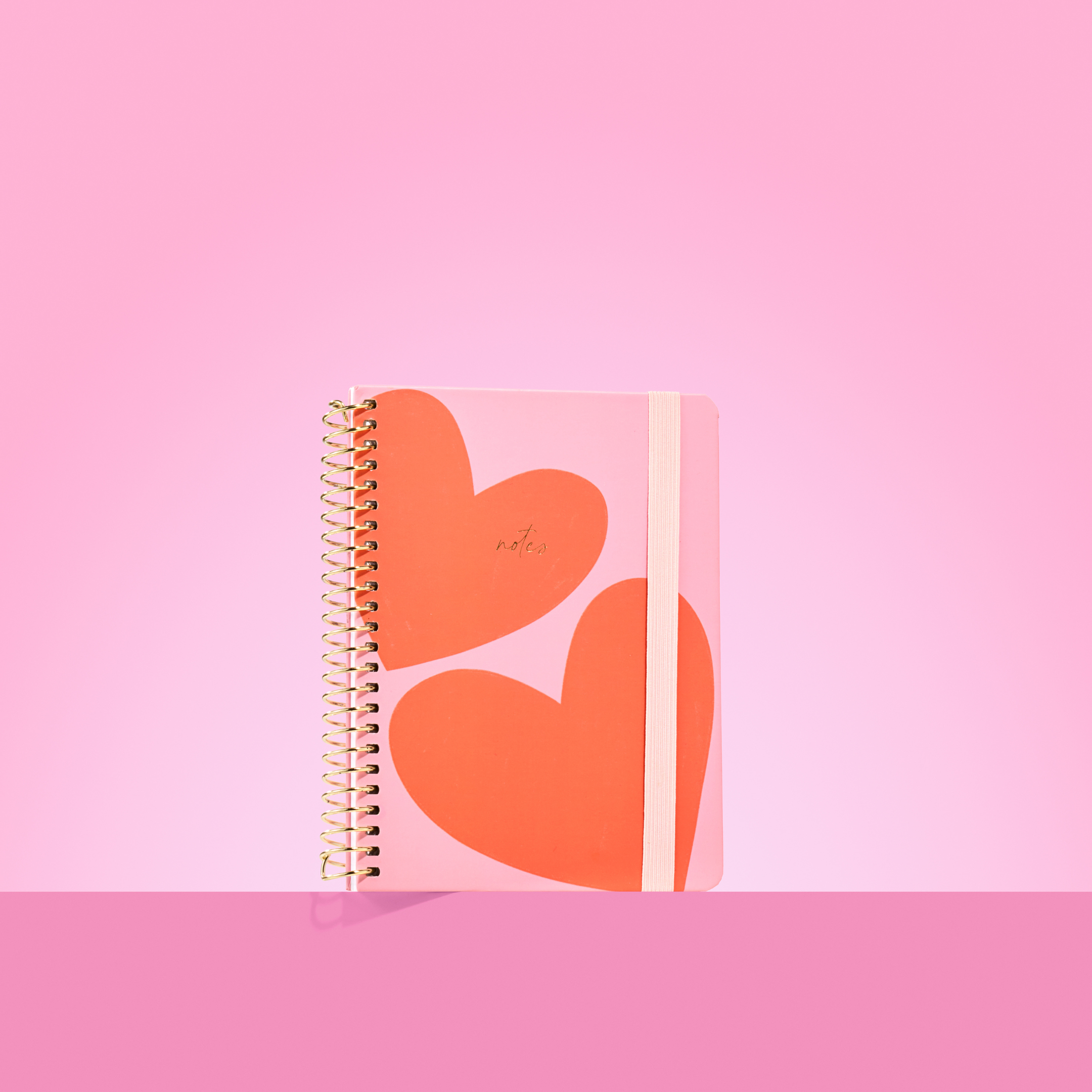 Pink 2026 notepad with two large red-orange hearts, gold spiral binding, light pink elastic closure, and "notes" embossed in gold.