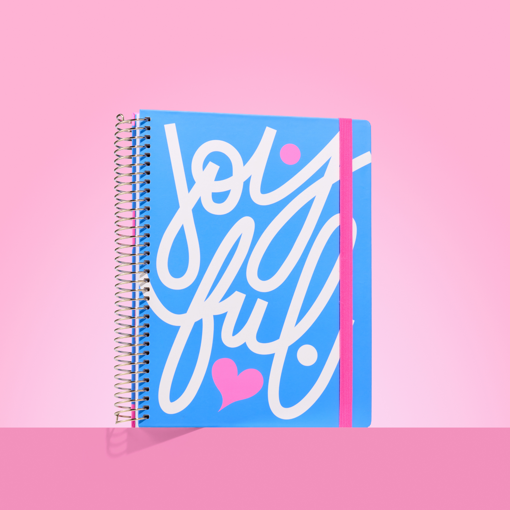 Blue 2026 spiral-bound planner with bold white "Joyful" script, pink heart detail, and pink elastic closure on a pink background.