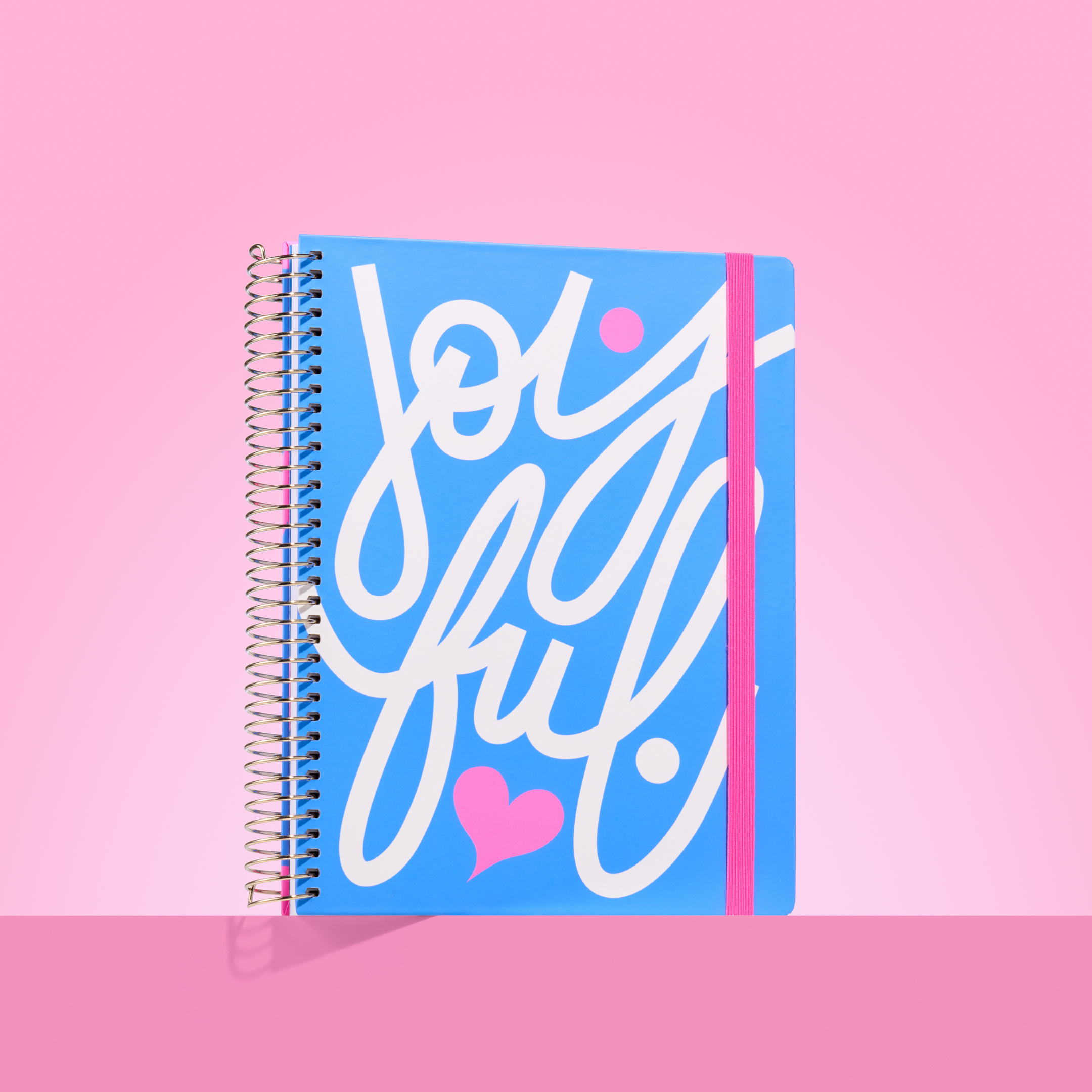 Blue 2026 spiral-bound planner with bold white "Joyful" script, pink heart detail, and pink elastic closure on a pink background.