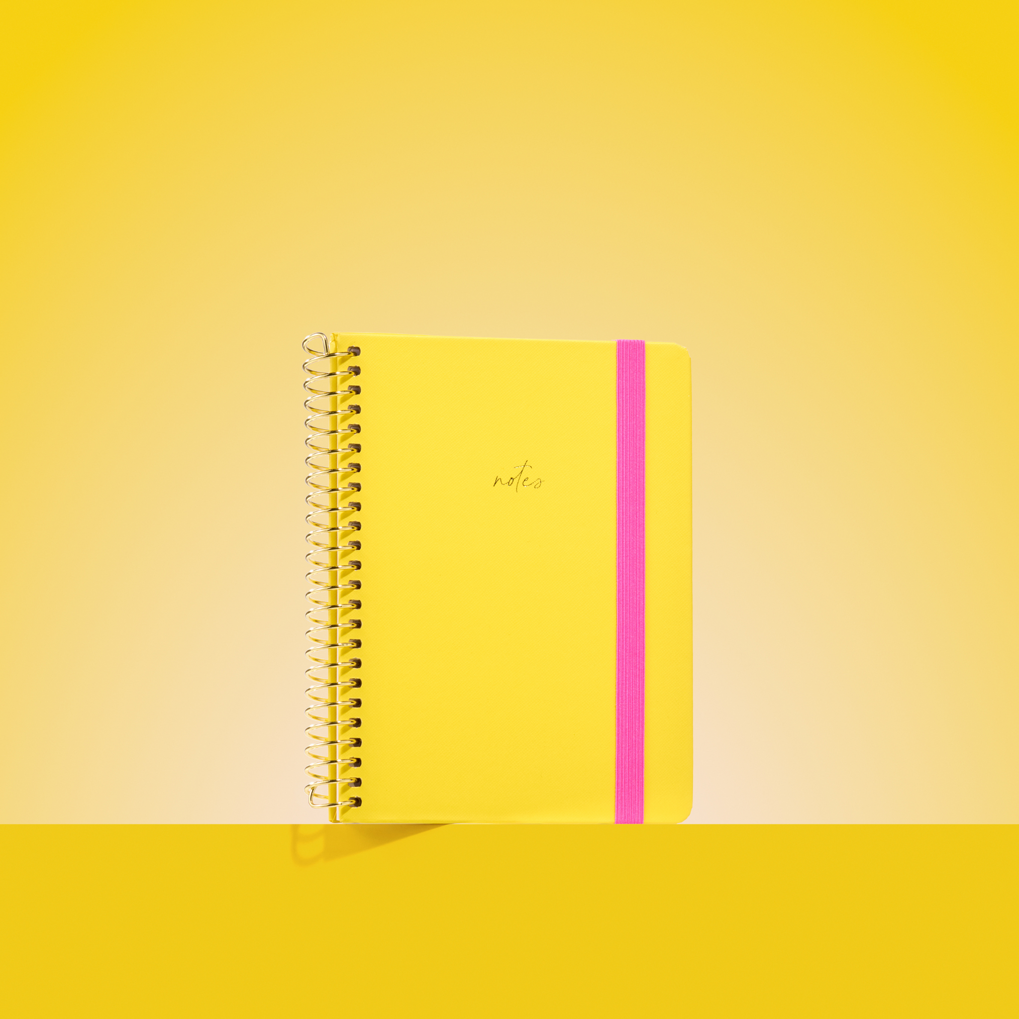 Bright yellow 2026 notepad with gold spiral binding, hot pink elastic closure, and "notes" embossed in gold on the cover.
