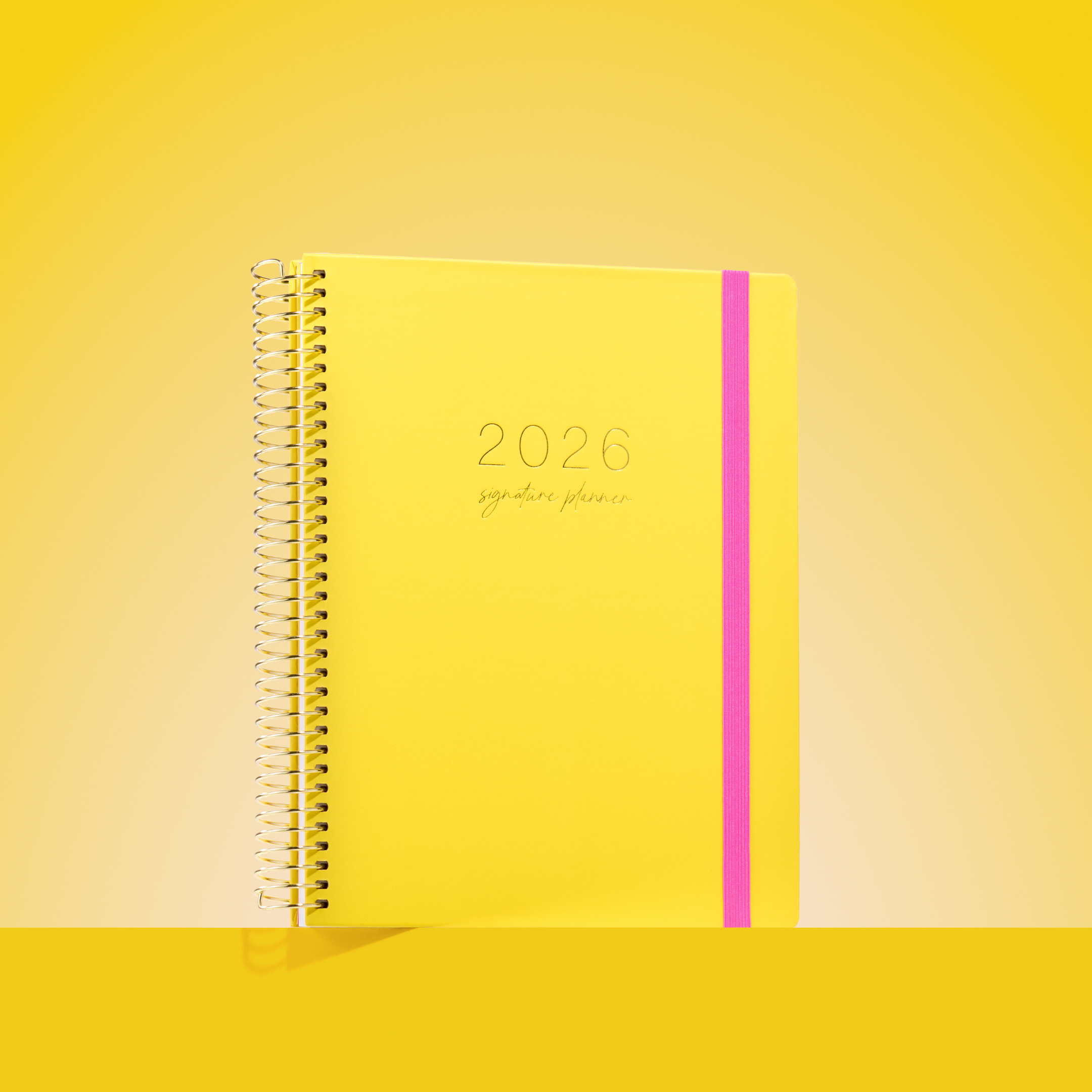 Bright yellow 2026 Signature Planner with hot pink elastic band, styled on a yellow background for a bold, modern look.