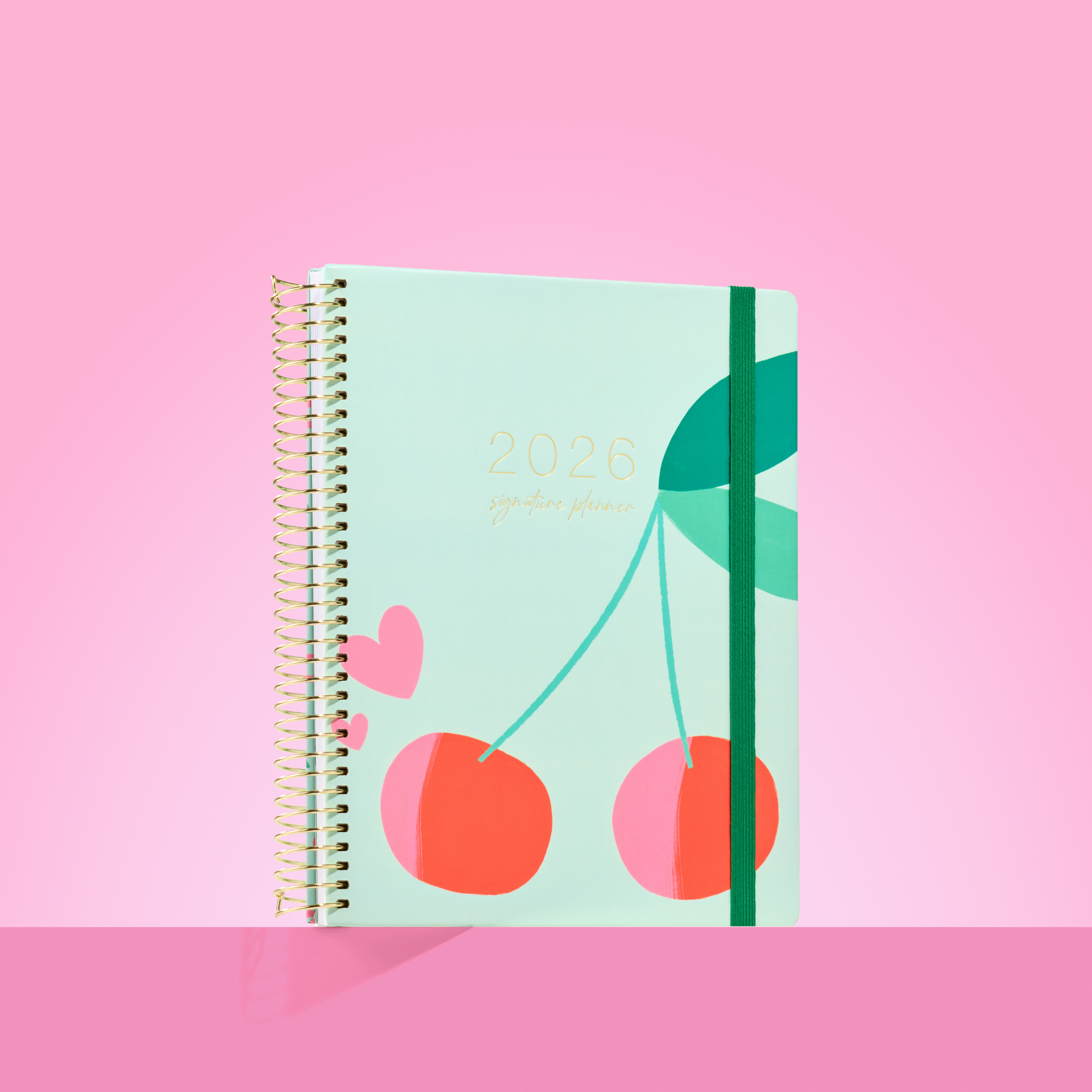 2026 teacher planner with bold cherry illustration on a mint cover, gold coil binding, and dark green elastic strap.
