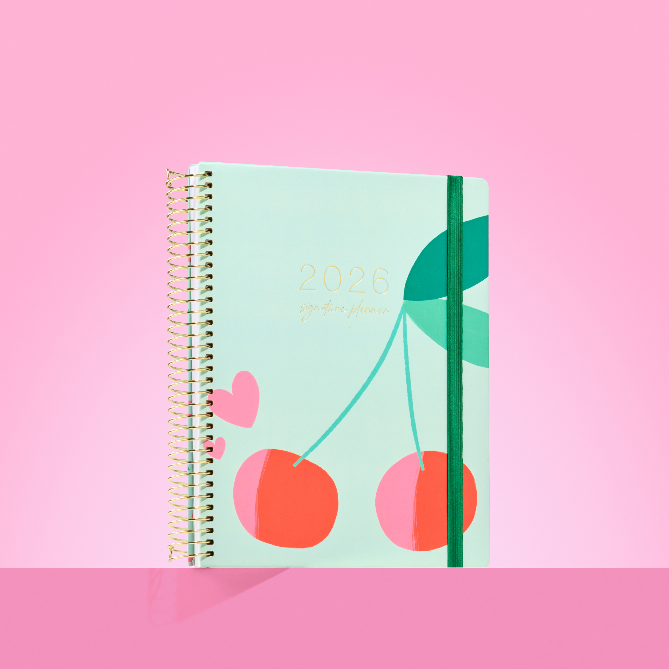 2026 teacher planner with bold cherry illustration on a mint cover, gold coil binding, and dark green elastic strap.