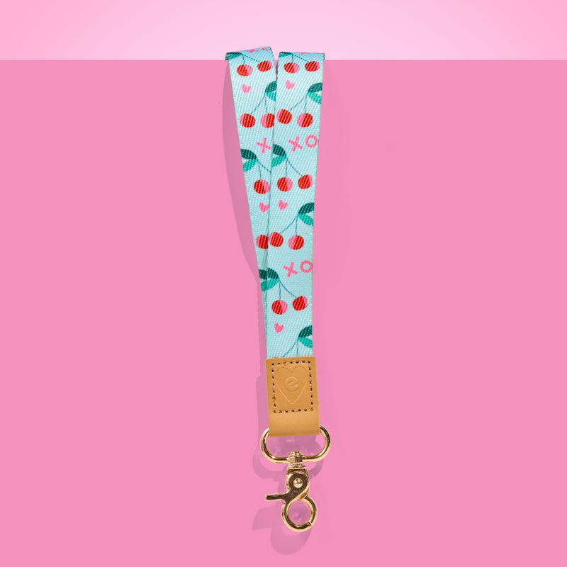 Cherry patterned teacher lanyard with tan leather accent and gold clasp, displayed on a bright pink background.
