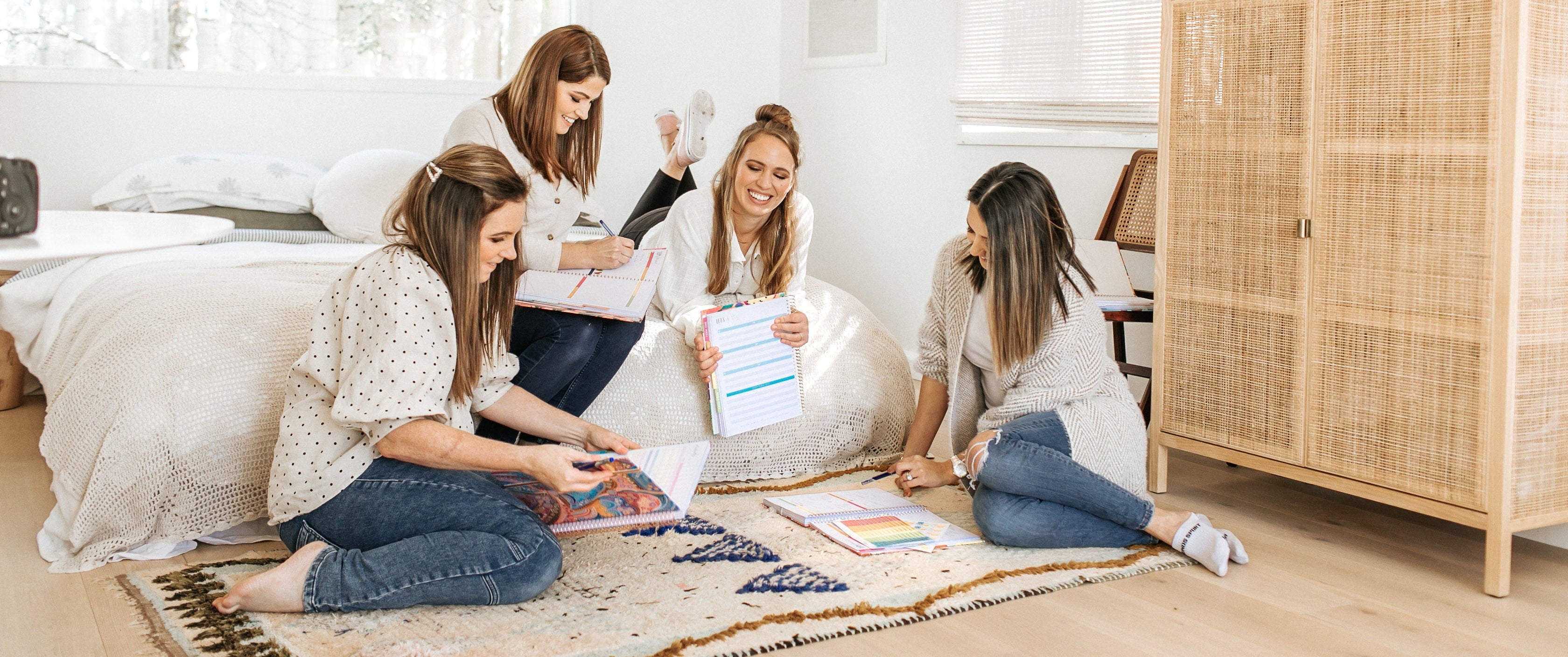Four women sitting on a rug in a bedroom with a wardrobe and window in the background.