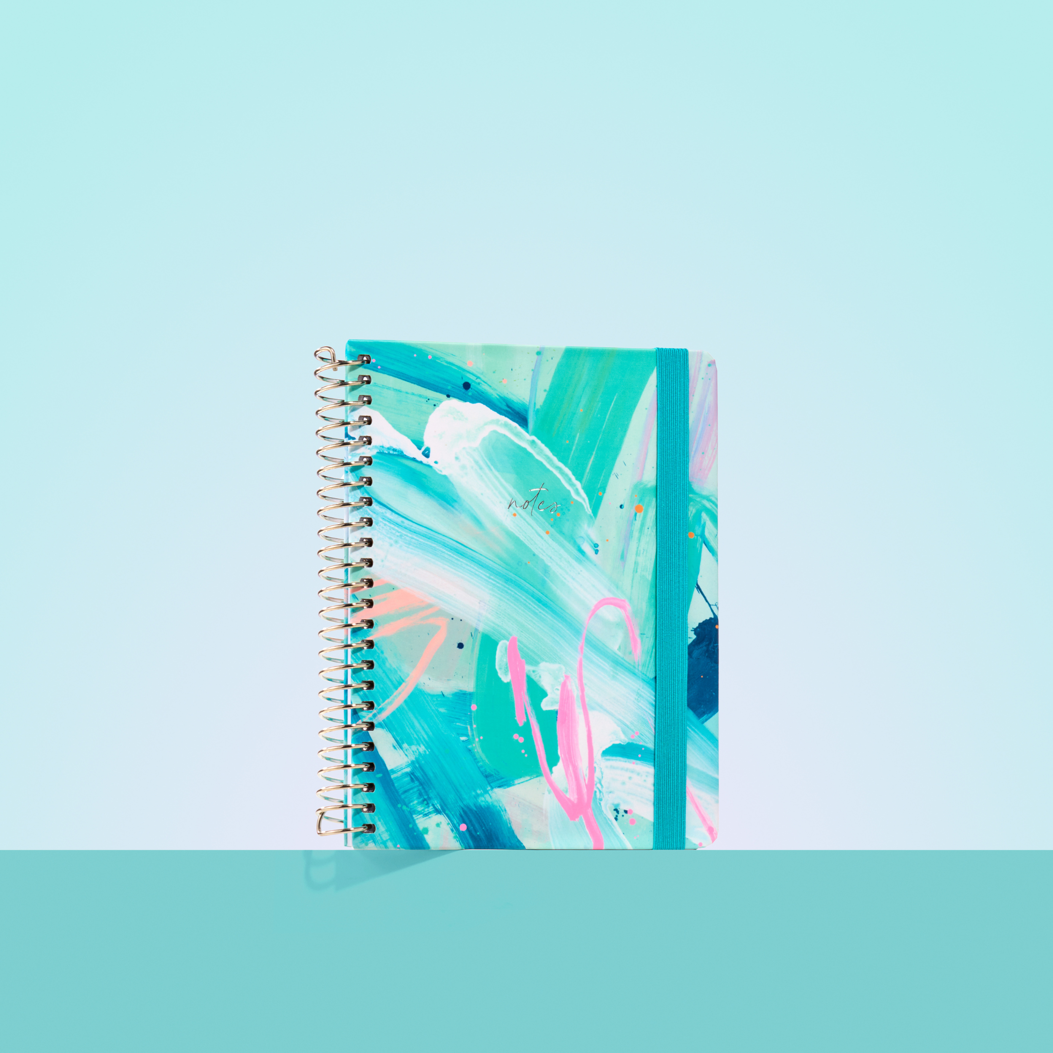 Teal and aqua 2026 notepad with abstract brushstroke design, silver spiral binding, dark teal elastic closure, and “notes” in gold.

