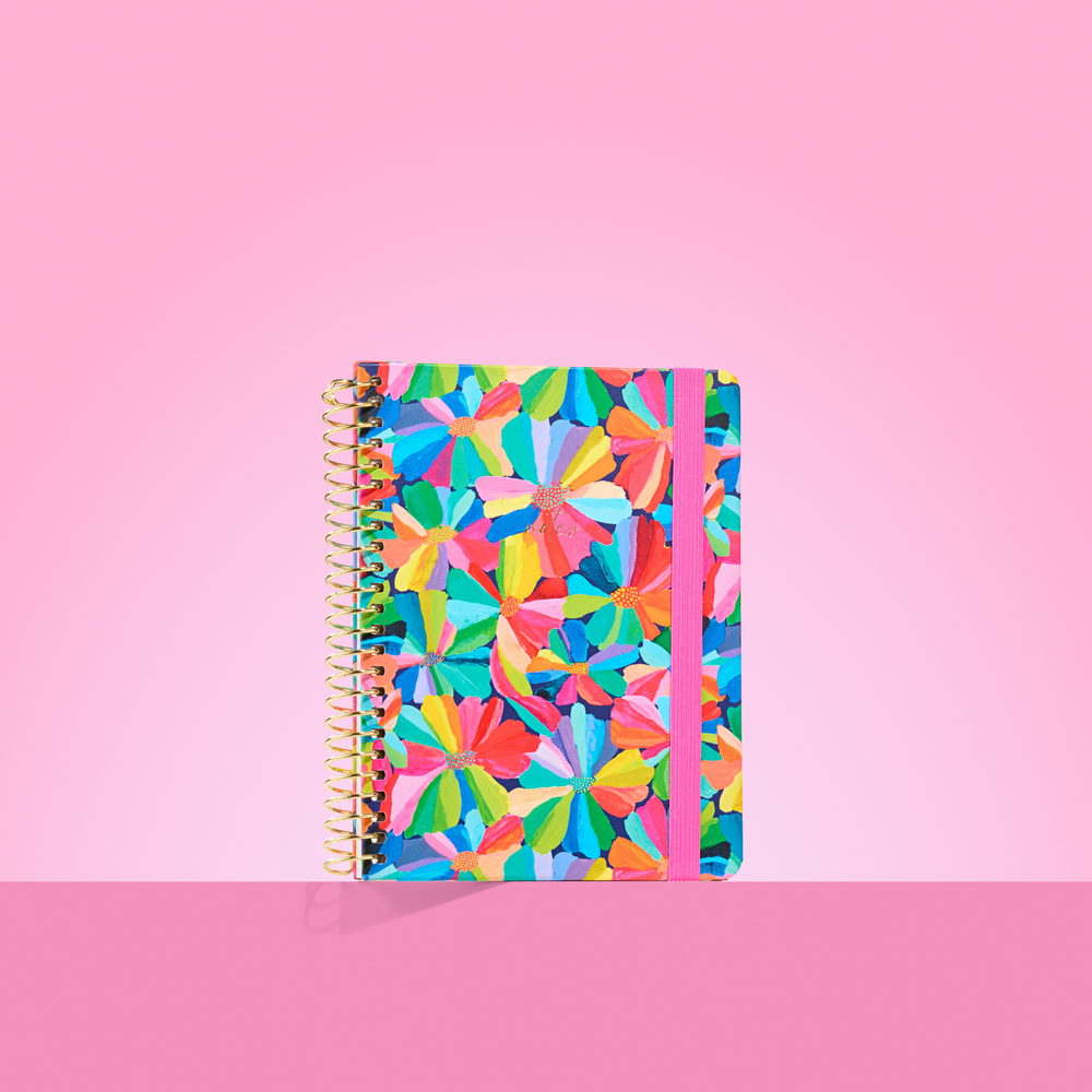 Colourful 2026 notepad with bold rainbow petal flower pattern, gold spiral binding, and pink elastic closure on a pink background.