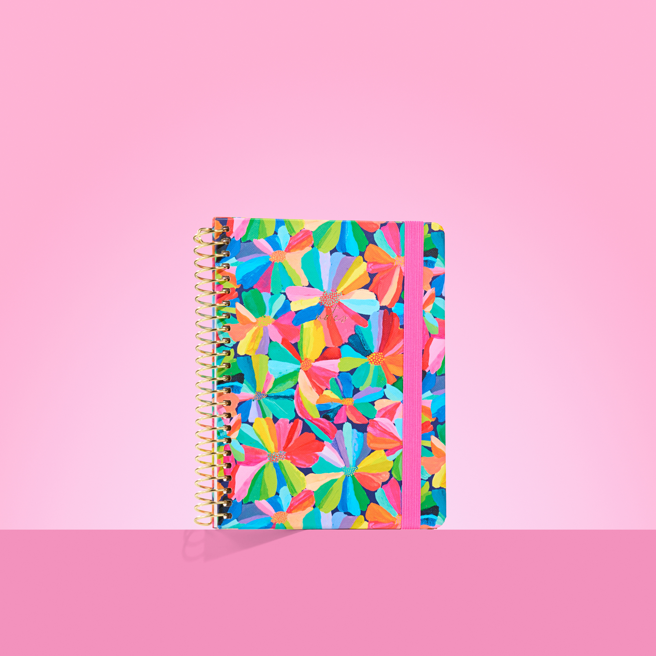 Colourful 2026 notepad with bold rainbow petal flower pattern, gold spiral binding, and pink elastic closure on a pink background.