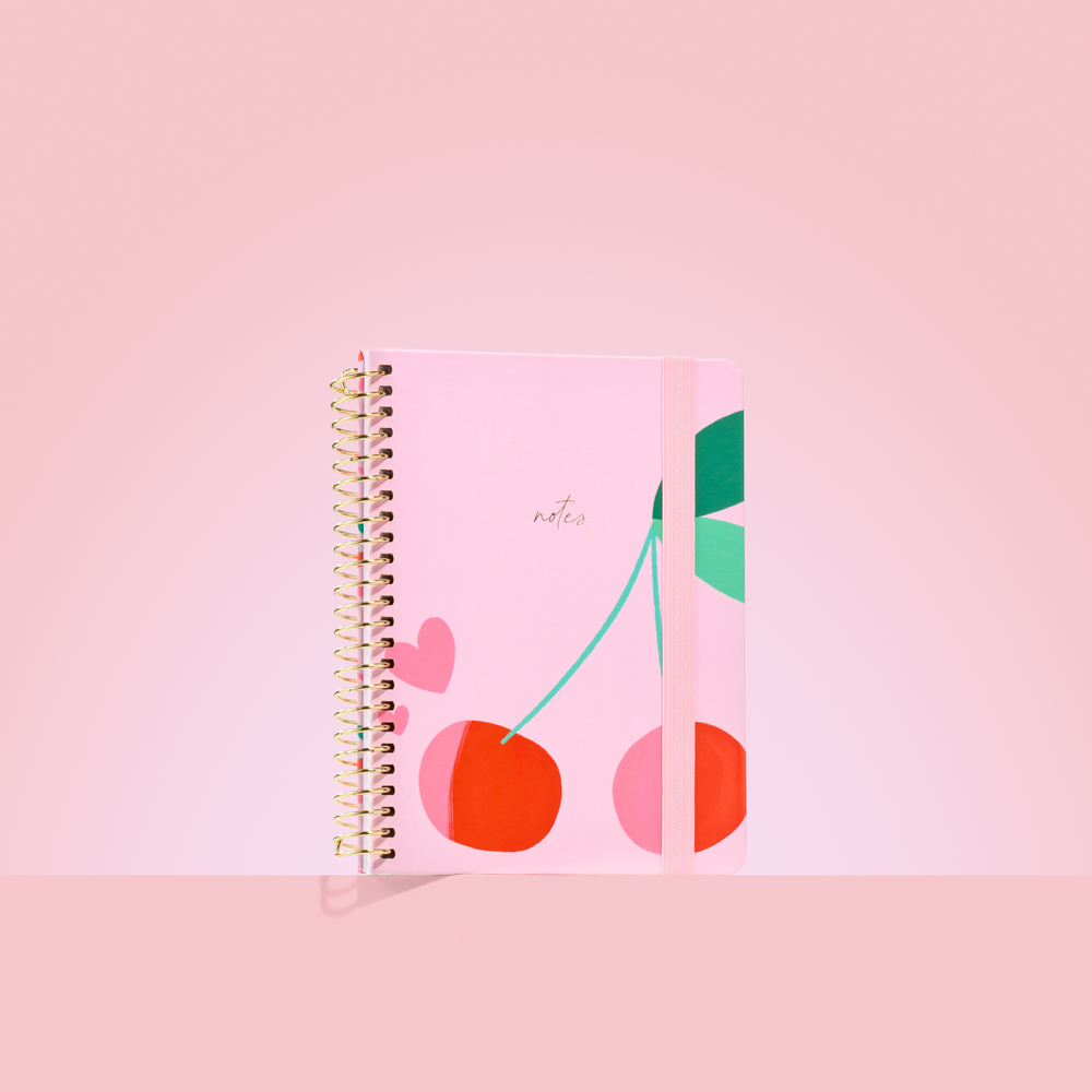 Pink 2026 notepad with red cherries and green leaves, gold spiral binding, elastic closure, and "notes" embossed in gold.