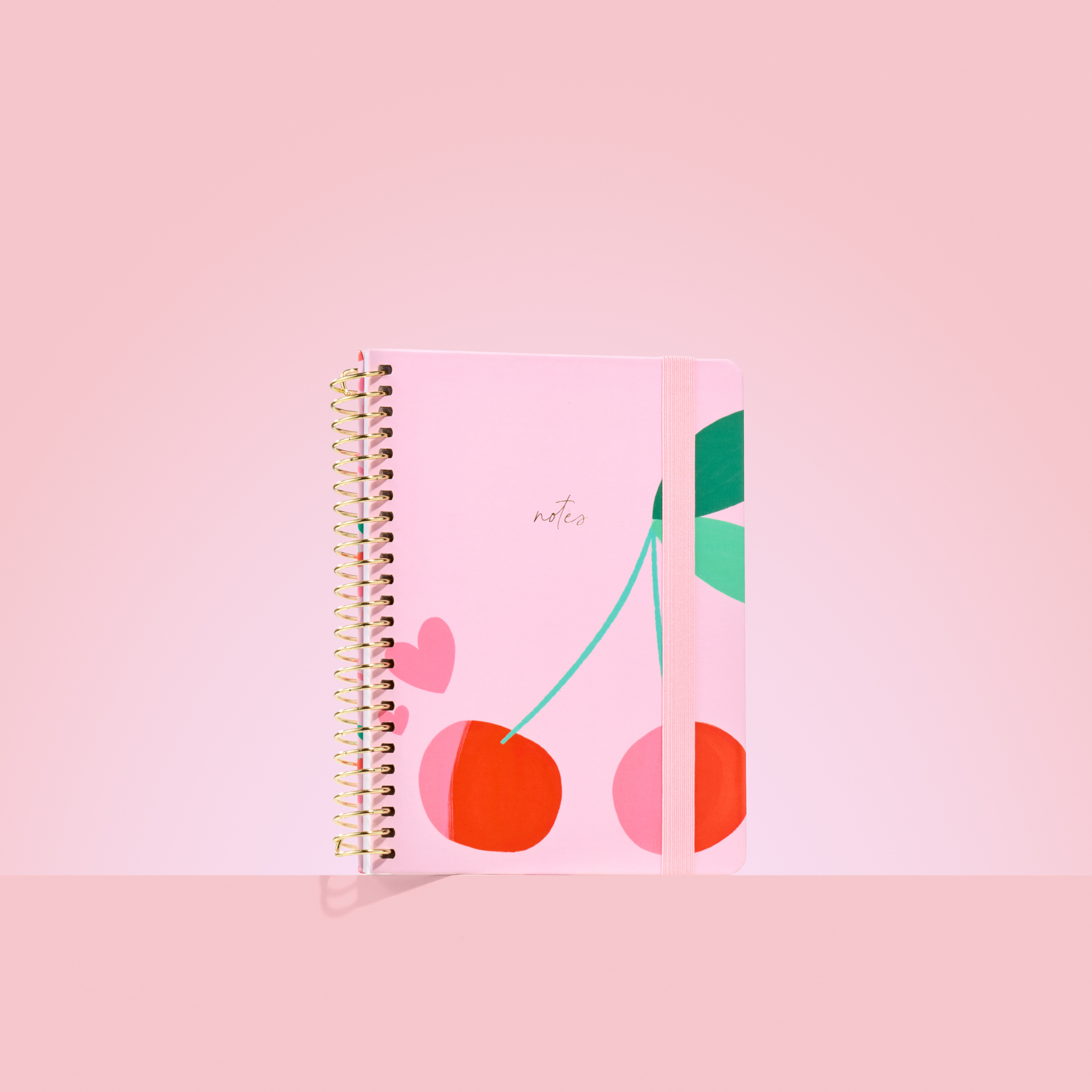 Pink 2026 notepad with red cherries and green leaves, gold spiral binding, elastic closure, and "notes" embossed in gold.