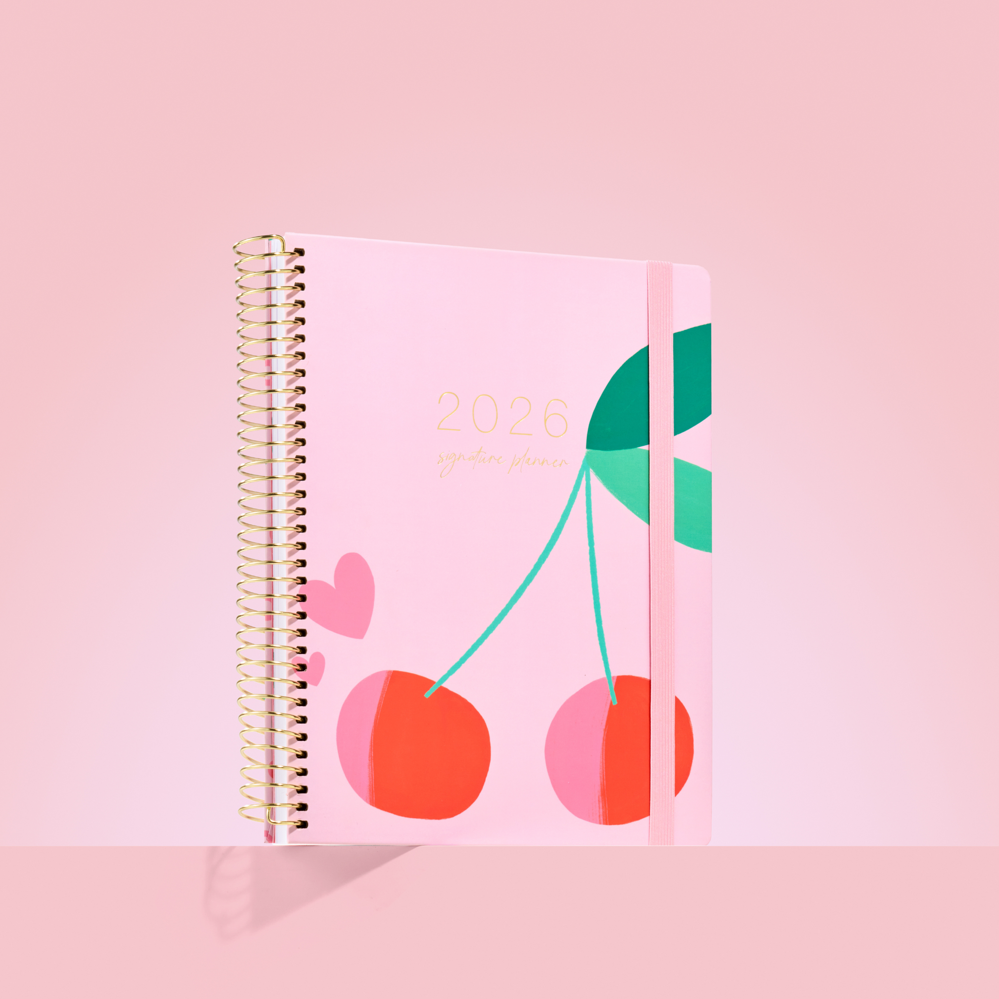 2026 planner with playful cherry illustration on a blush pink background, gold coil binding, and pink elastic strap.

