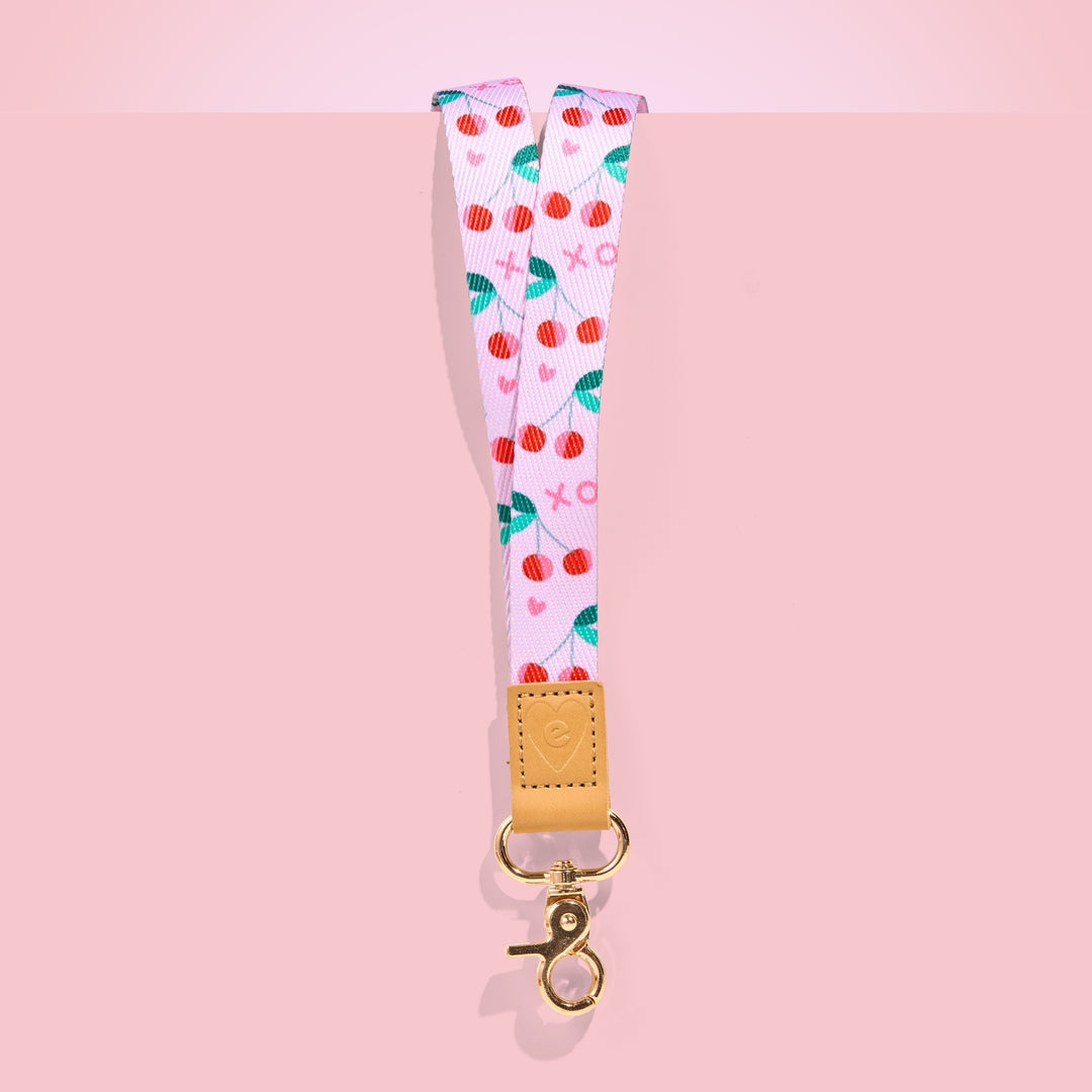 Pink teacher lanyard with red cherry pattern, tan leather accent and gold clasp, displayed on a blush pink background.