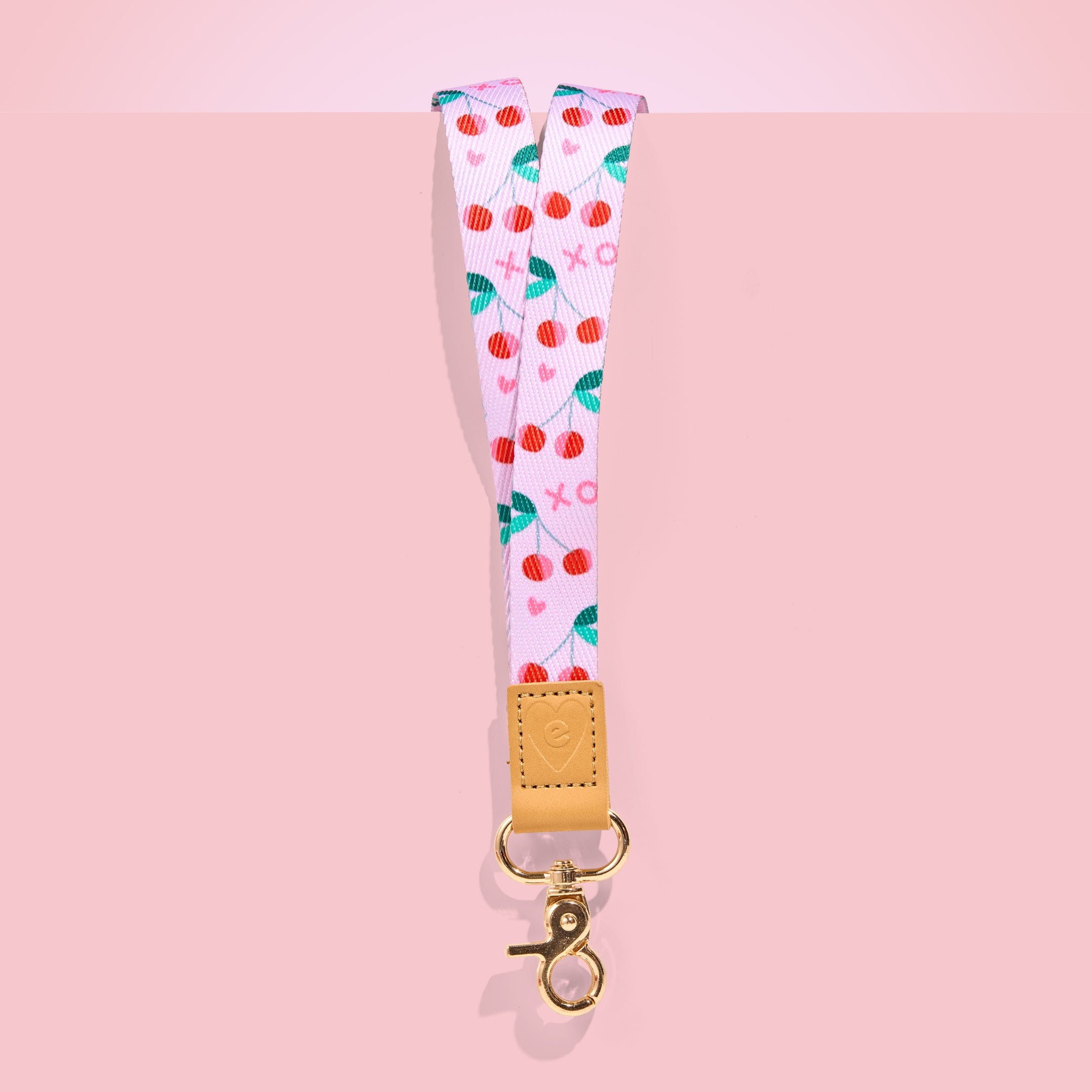 Pink teacher lanyard with red cherry pattern, tan leather accent and gold clasp, displayed on a blush pink background.