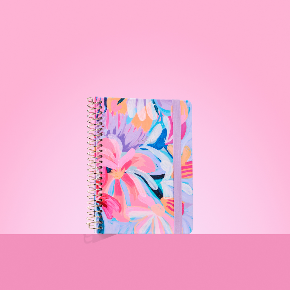 Colourful floral 2026 notepad with spiral binding and pink elastic closure, set against a two-tone pink background.

