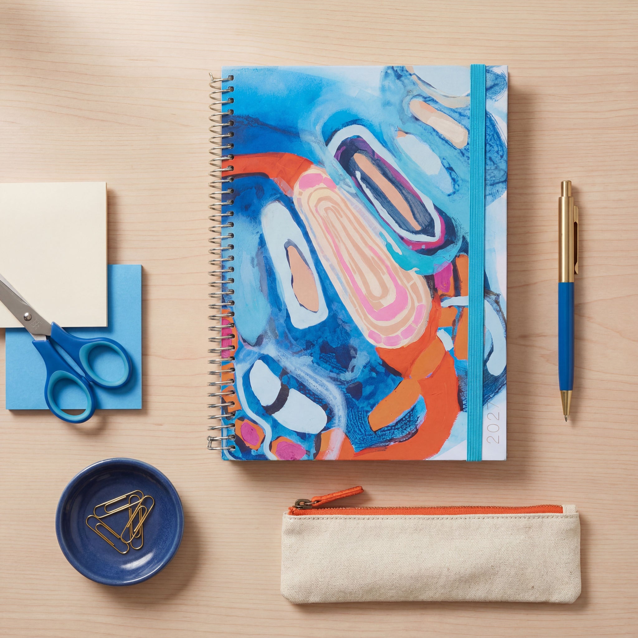 2027 slimline abstract art spiral planner styled on a light wood desk with blue scissors, gold pen, paperclips and a pencil case.