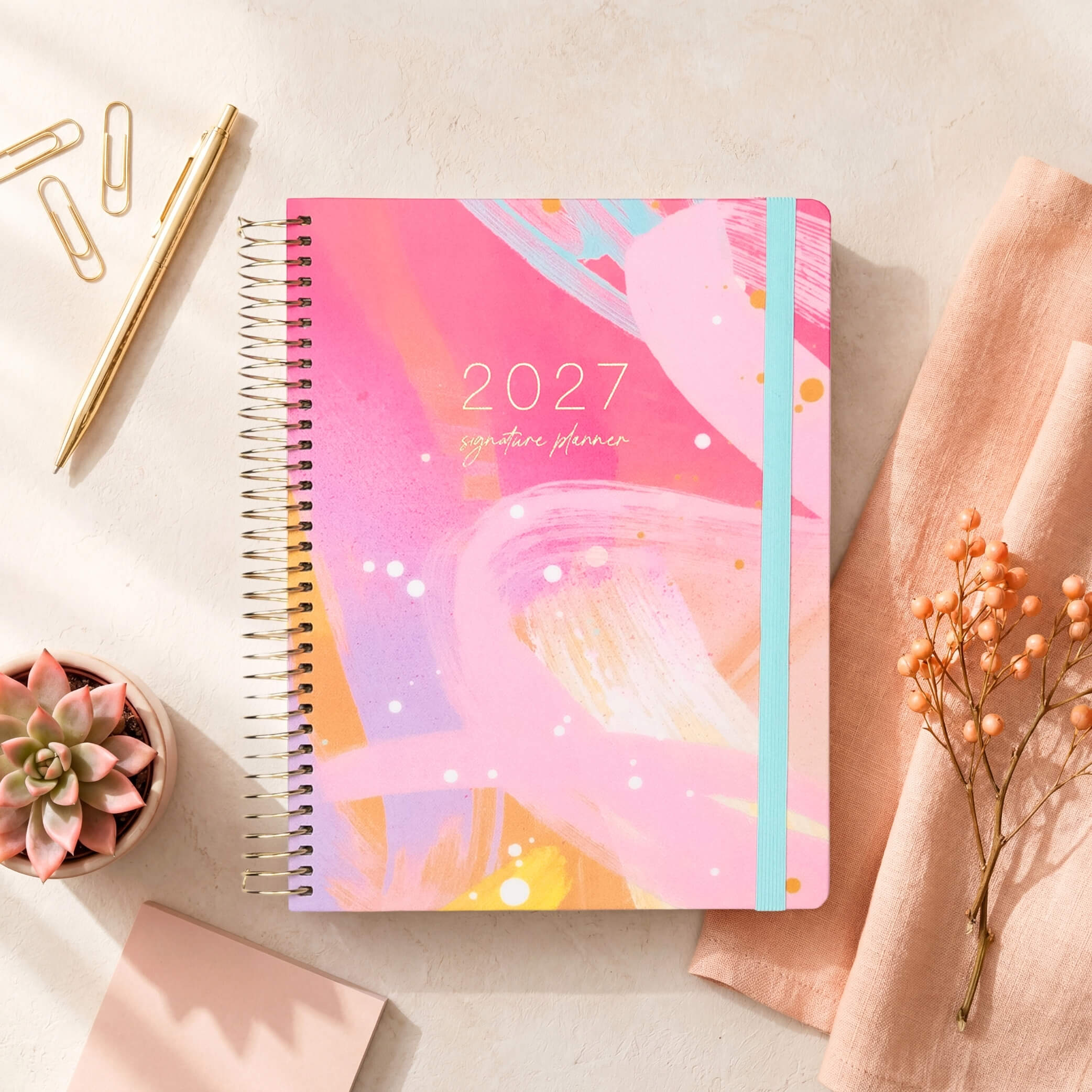 2027 Signature Planner in Solar Glow styled on a cream surface with a succulent and linen, gold coil and teal elastic closure.