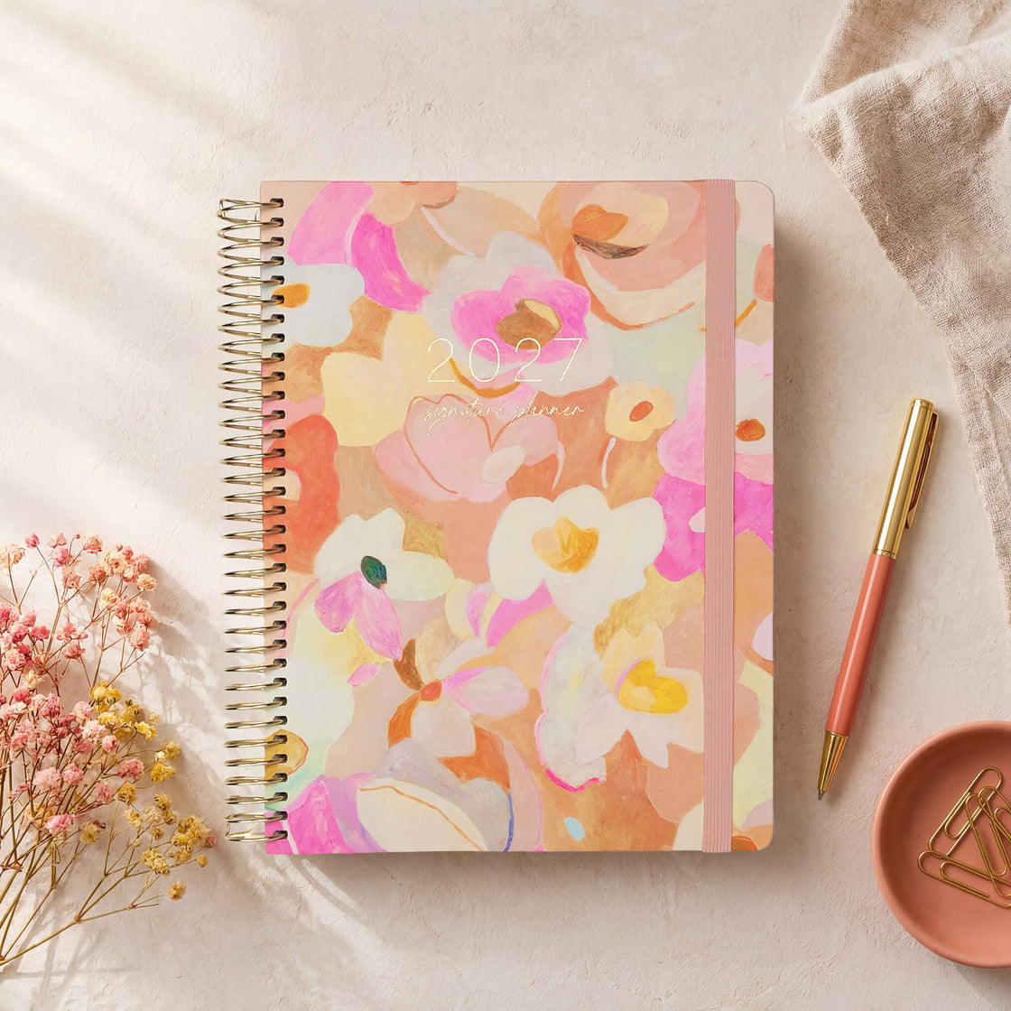 2027 Signature Planner in Love Fool styled with dried florals and a pink pen, gold coil and pink elastic closure.