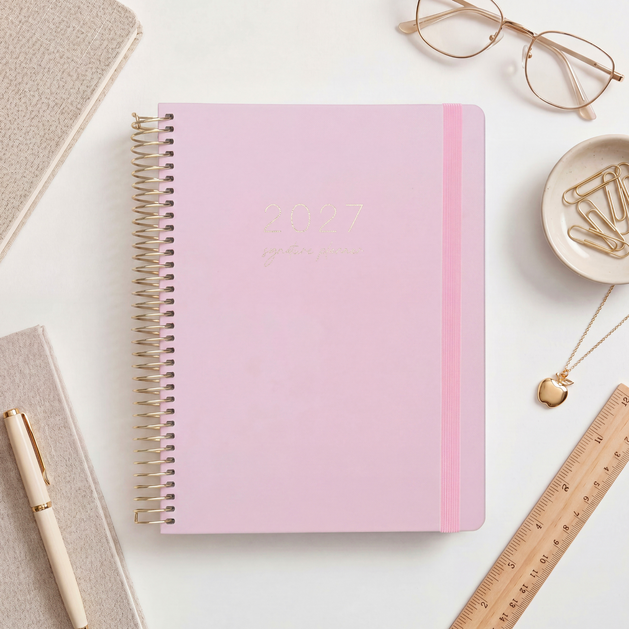 2027 Signature Planner in Blush styled with gold glasses and a wooden ruler, gold coil and pink elastic closure.