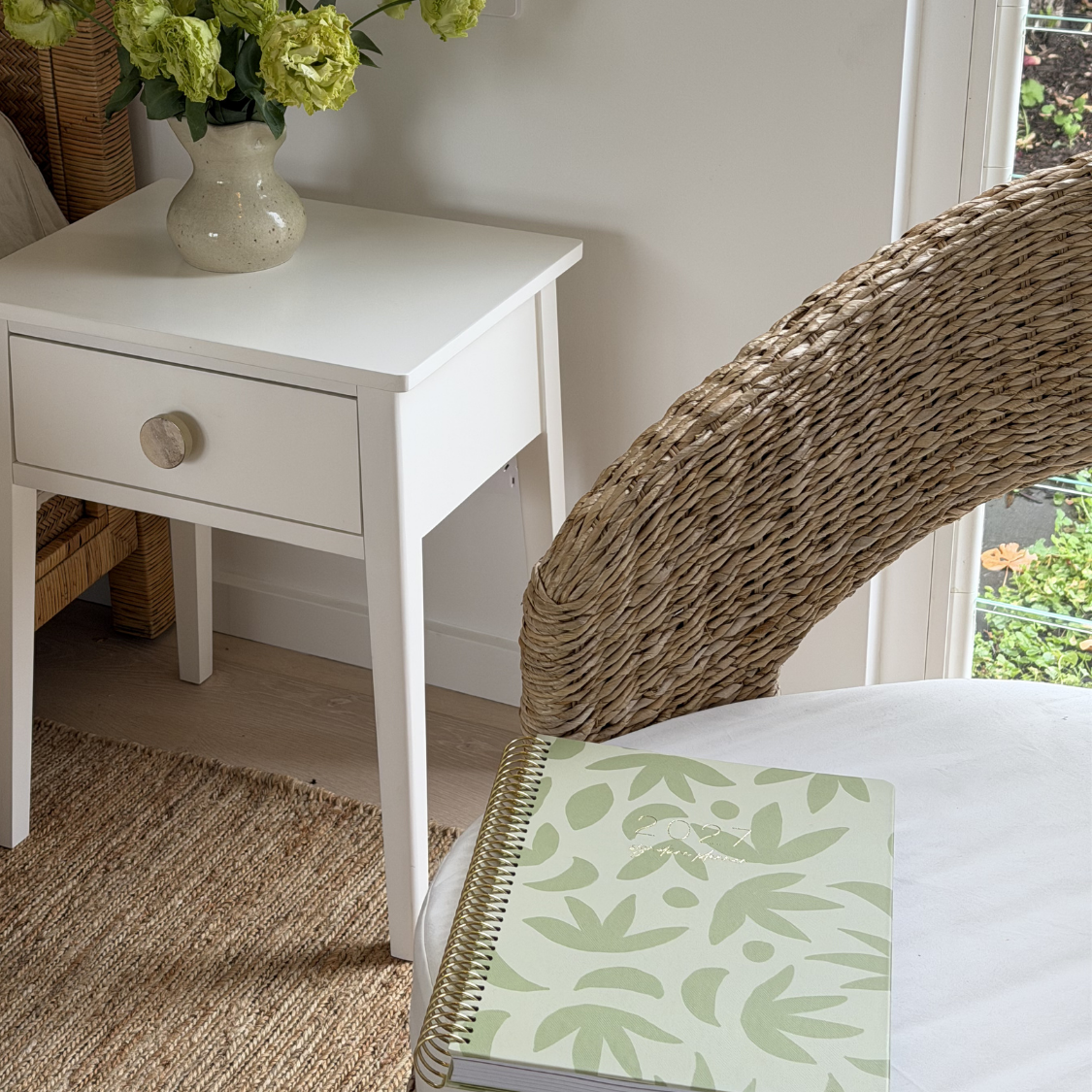 2027 Signature Planner in Sunburst Sage resting on a white chair in a styled living room with green florals, gold coil.