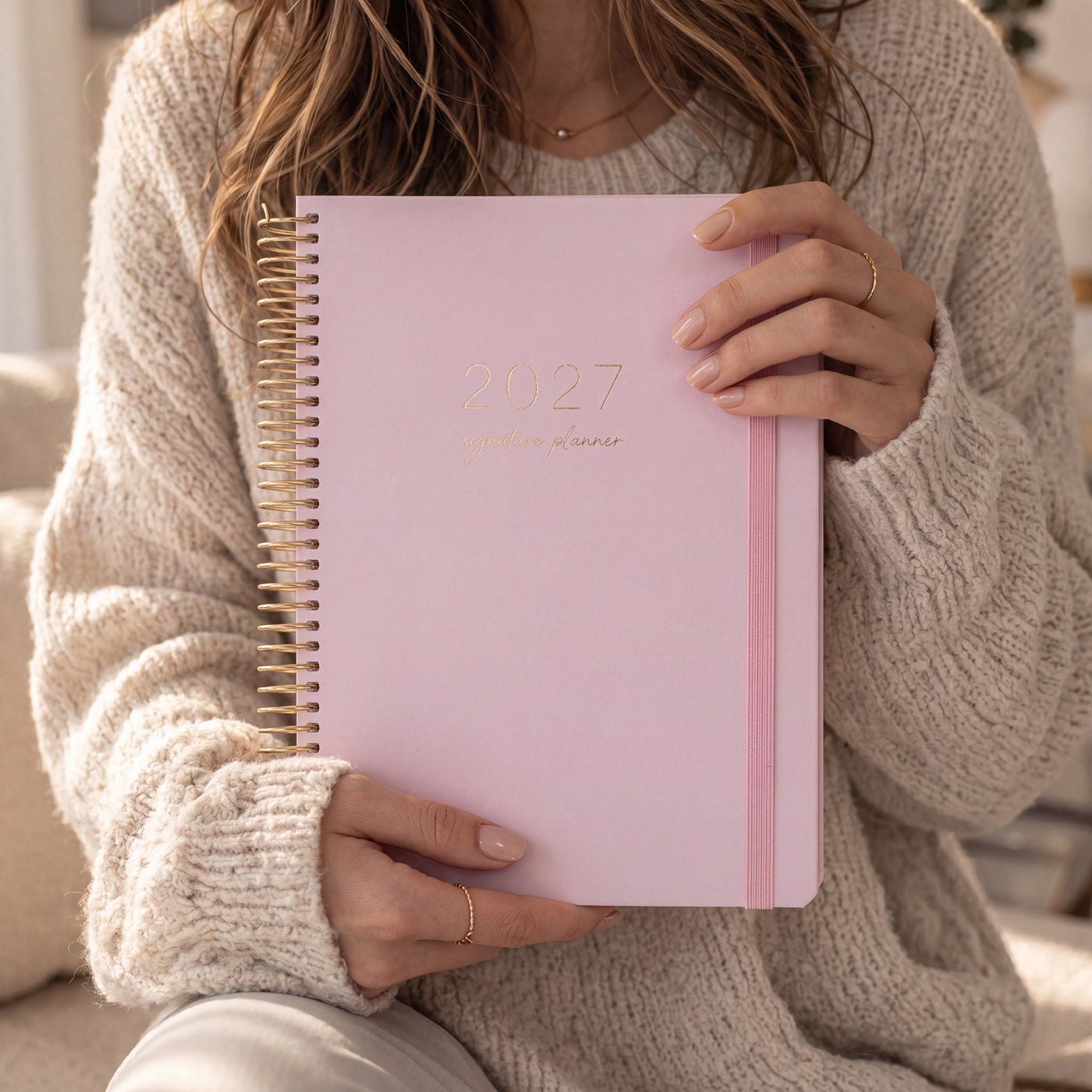 2027 Signature Planner in Blush held by a woman in a cream knit sweater, gold foil text, gold coil and pink elastic closure.