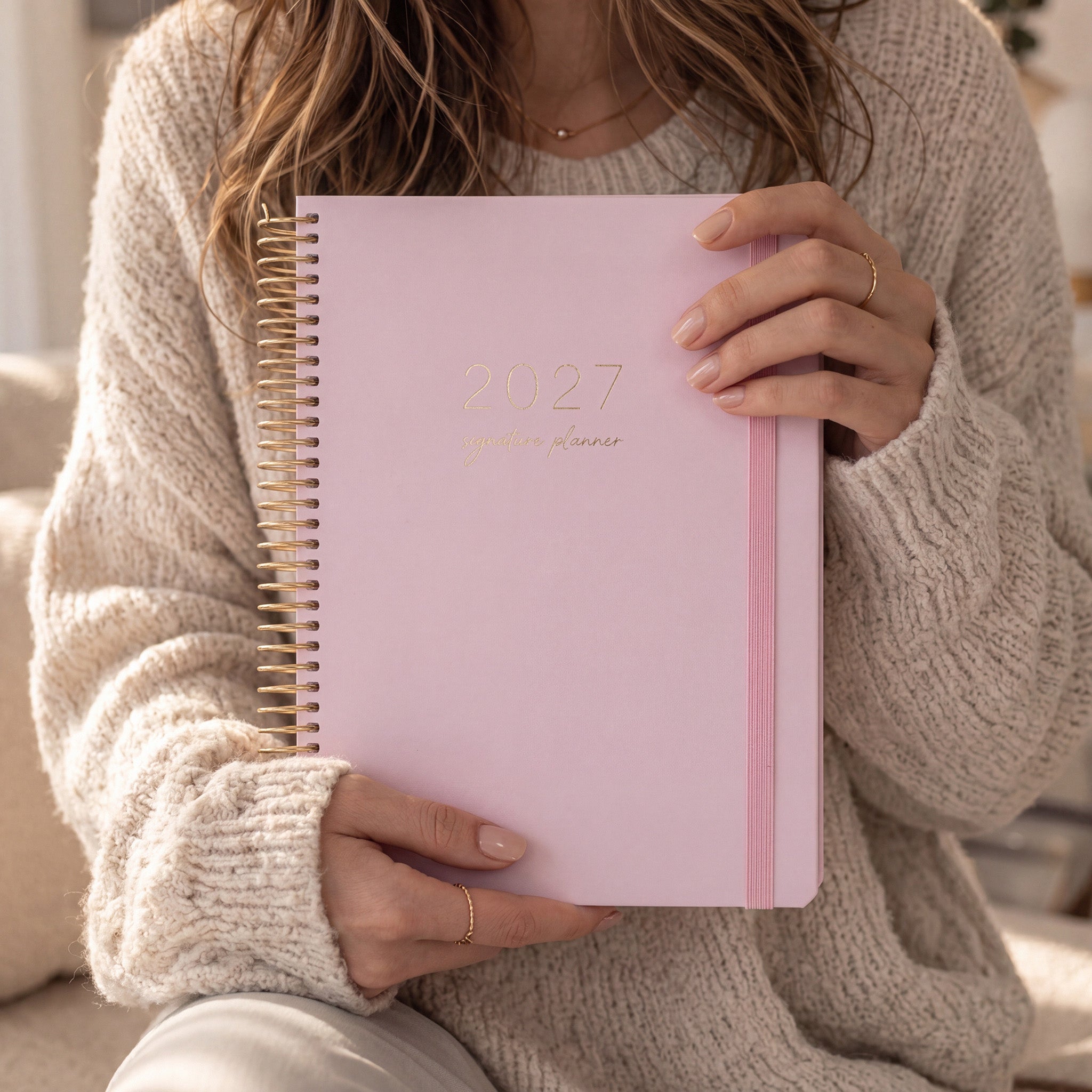 2027 Signature Planner in Blush held by a woman in a cream knit sweater, gold foil text, gold coil and pink elastic closure.