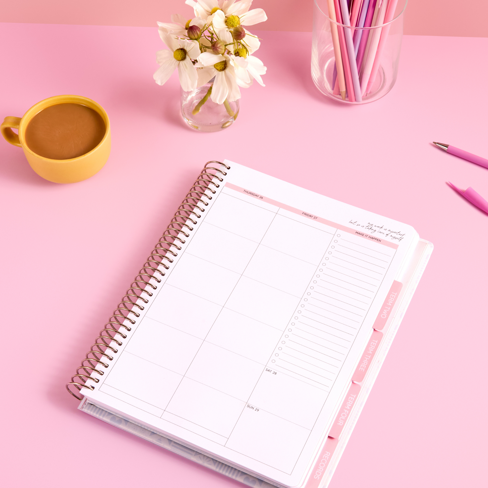 Open 2026 planner showing weekly layout, pastel pink pens, white flowers, and a yellow mug on pink desk.