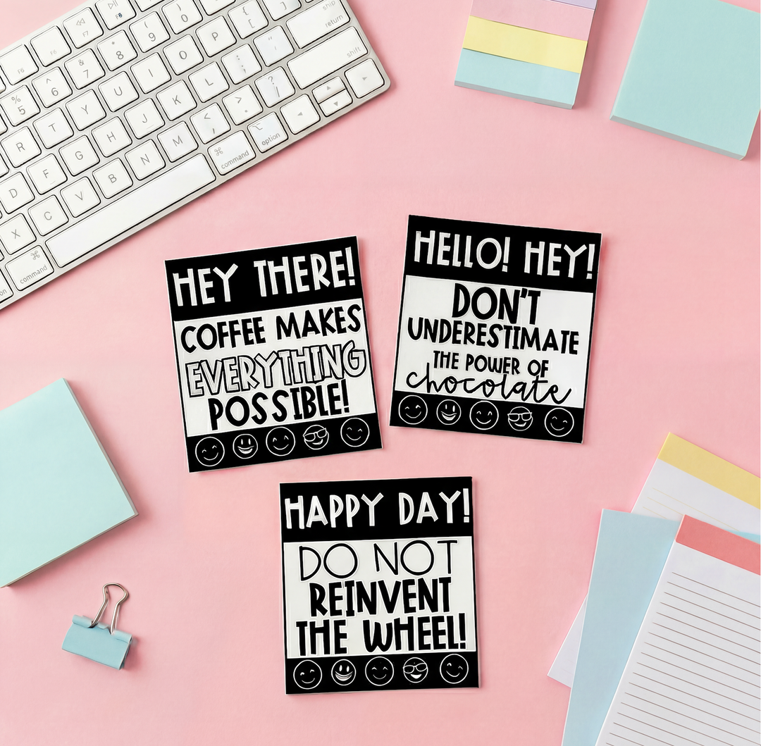 Three black-and-white motivational note cards with playful messages about coffee, chocolate, and positivity, arranged on a pink desk with a keyboard, sticky notes, and stationery items