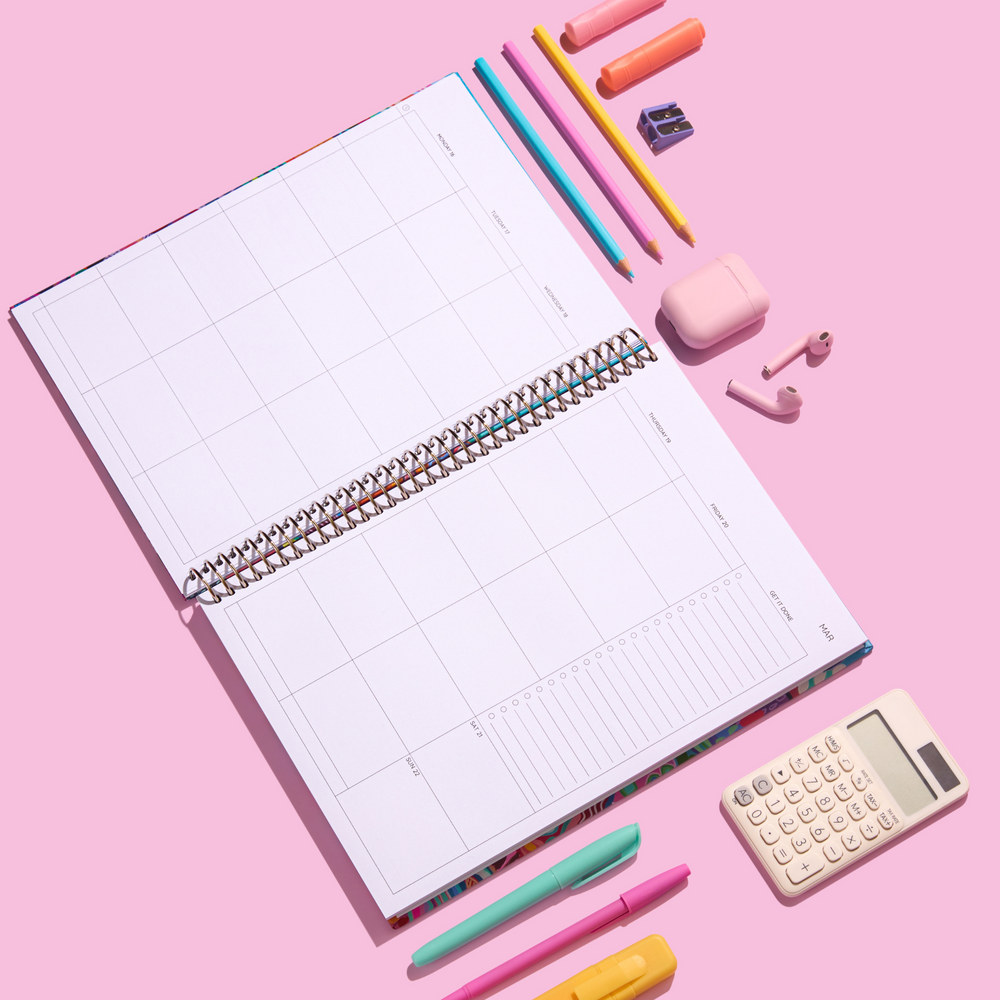 Flat lay of an open 2026 teacher planner showing a full weekly layout with accessories including pens, pencils, AirPods, highlighters, and a calculator on a pink background.