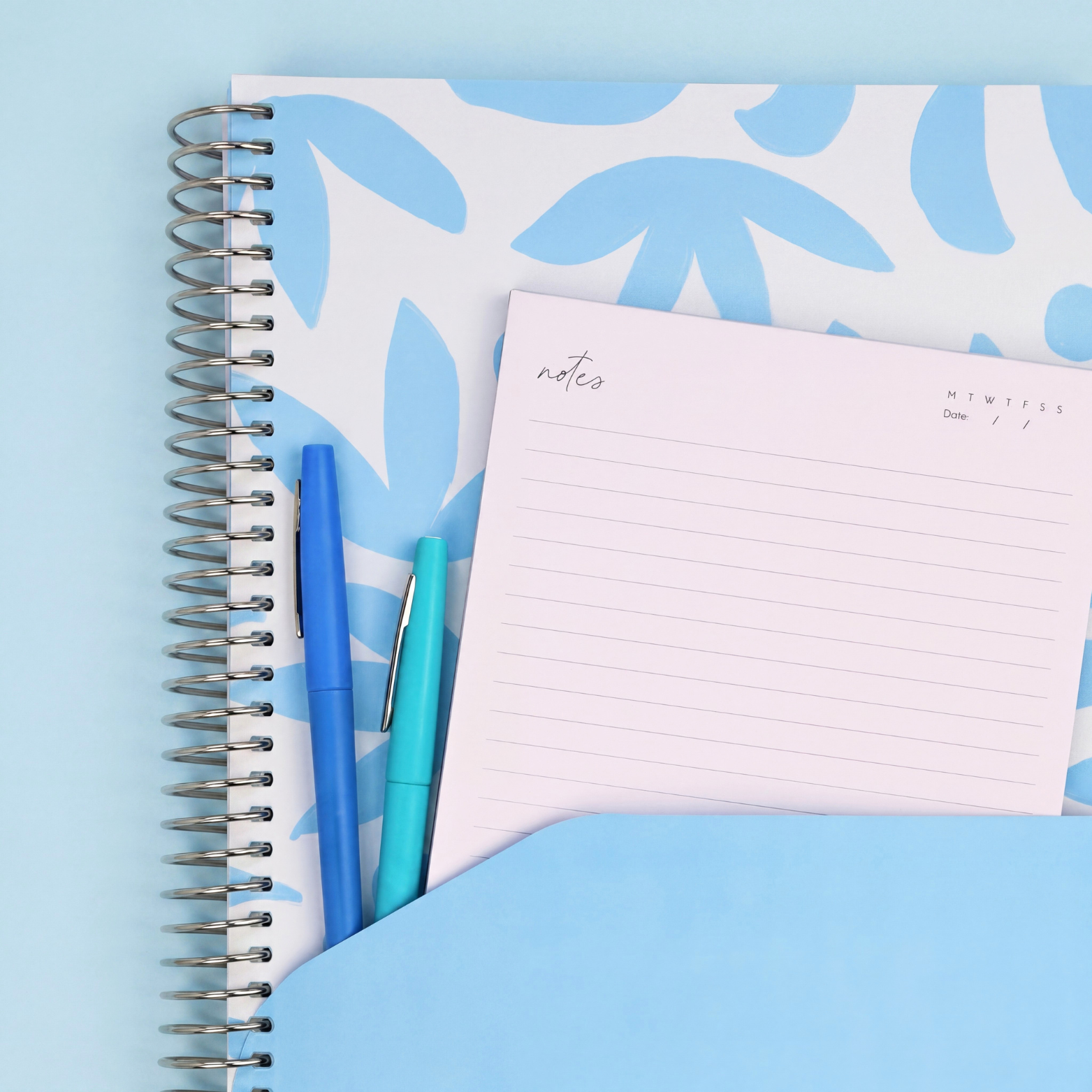 2027 Sunburst Sky Weekly Teacher Planner showing blue pocket sleeve with notes, pens and silver coil on a white background.