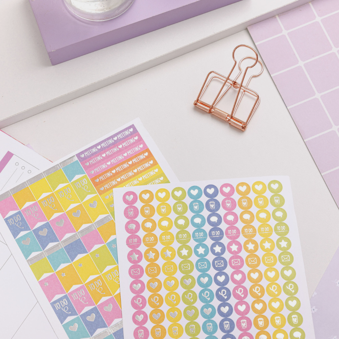 Two sheets of colourful planner stickers with icons for tasks, emails, meetings, and coffee on a pastel desk setup.