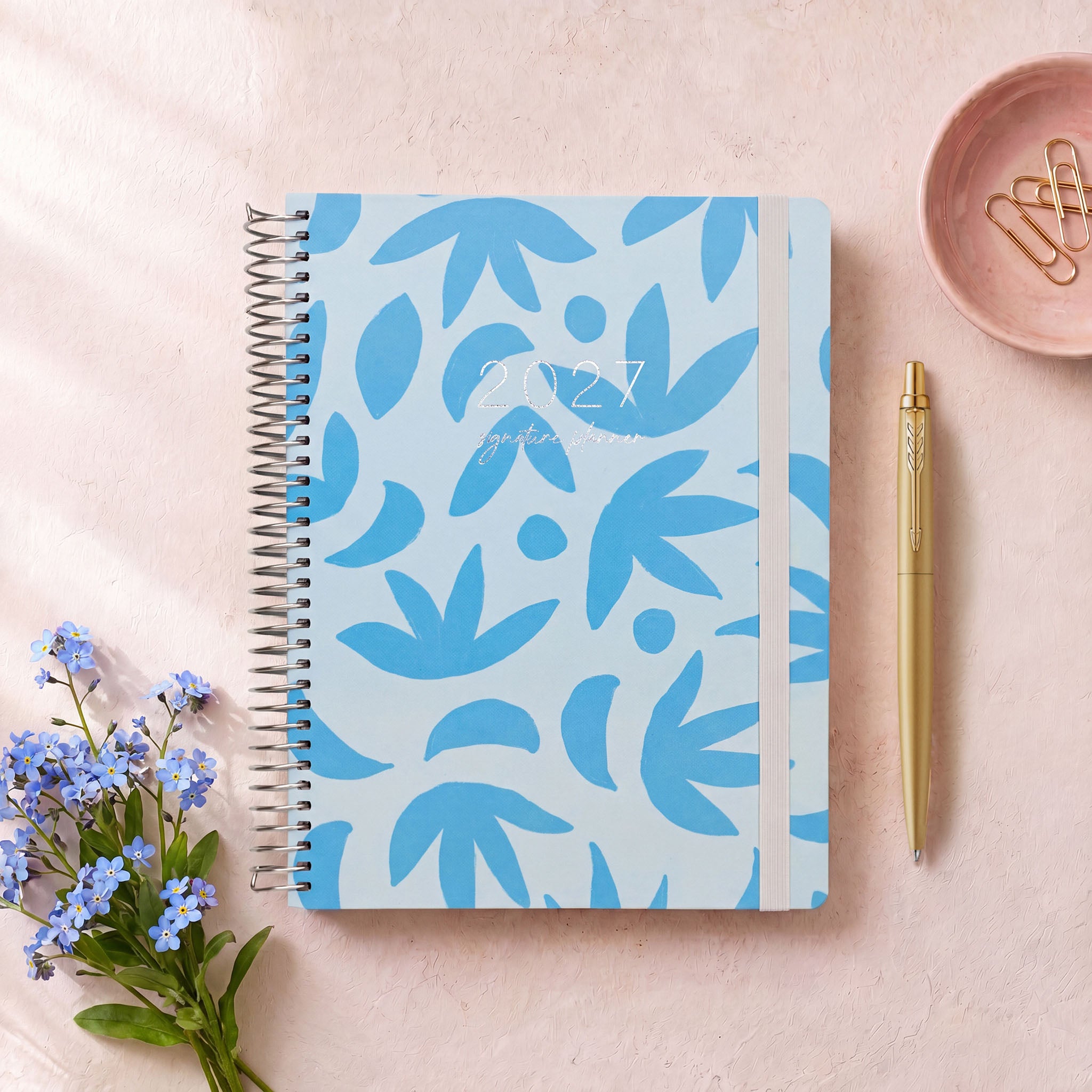 2027 Signature Planner in Sunburst Sky with blue leaf pattern, styled with blue flowers, silver coil and grey elastic closure.