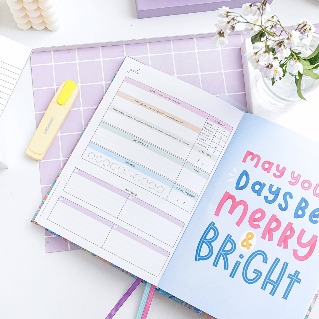 Open weekly planner lying on a desk, displaying a goals page on the left and large text on the right saying 'May your days be merry & bright.'