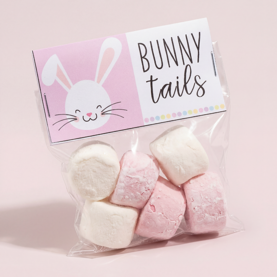 Close-up photo of a small clear treat bag filled with pink and white marshmallows labeled “BUNNY tails.” The topper card is pastel pink and white with a cute bunny face illustration on the left and playful handwritten-style text on the right. The bag is sealed with staples and displayed against a soft pink background.