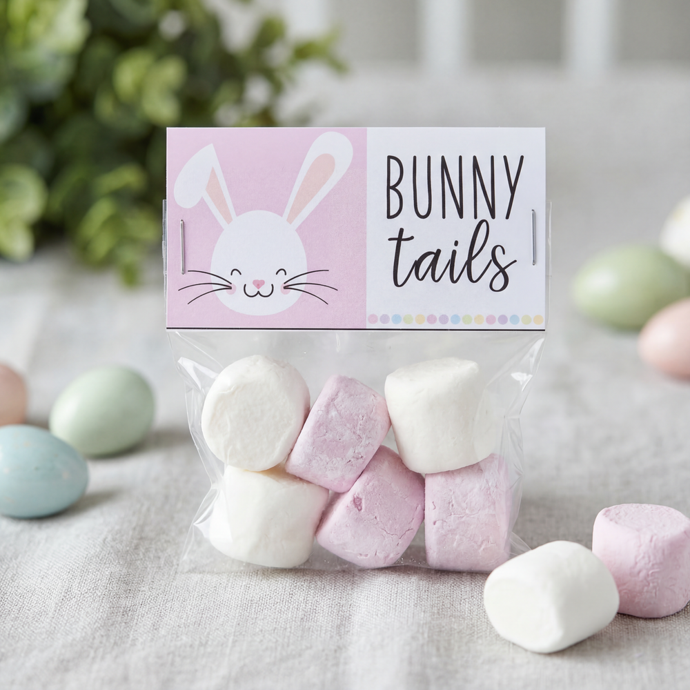A small clear gift bag filled with pink and white marshmallows sits on a soft fabric surface. The bag is topped with a pastel pink and white printable label featuring a smiling bunny face and the words “BUNNY tails” in playful lettering. Pastel Easter eggs and blurred greenery decorate the background, creating a soft spring-themed scene.