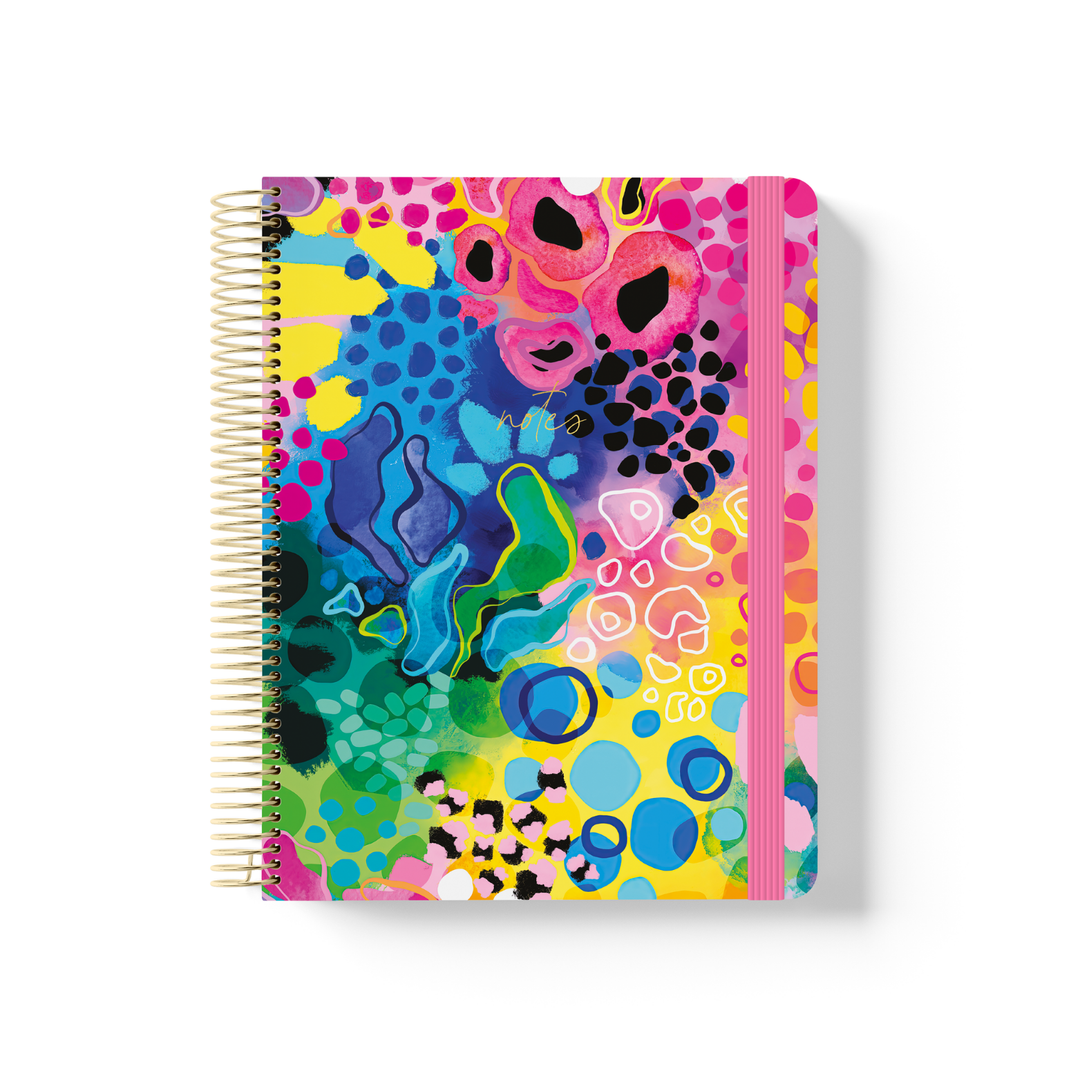 Boom spiral notebook with bold abstract artwork in pink, yellow, blue and green, gold coil binding, and pink elastic closure.