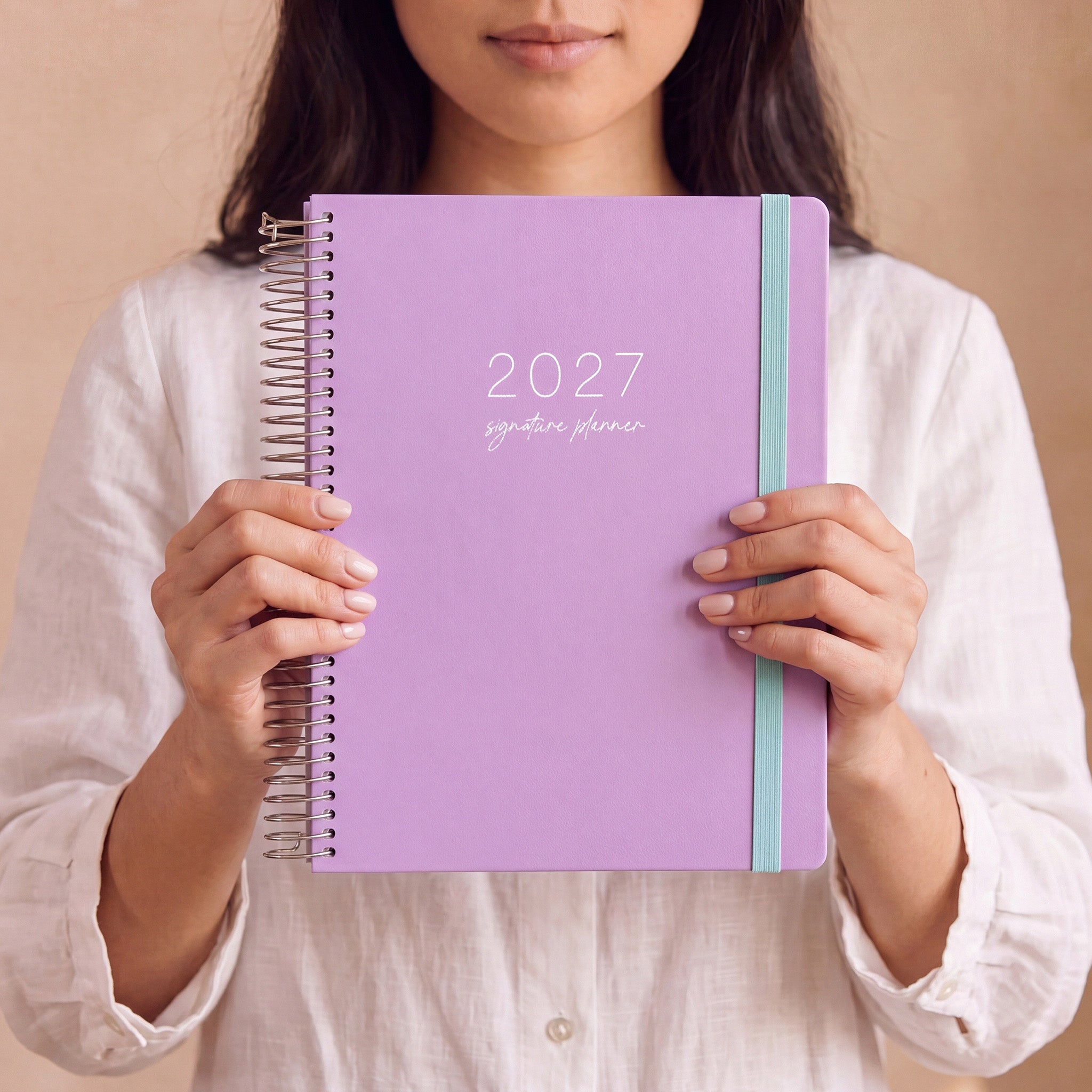 2027 Signature Planner in Lilac held by a woman in a white linen shirt, silver coil and teal elastic closure.