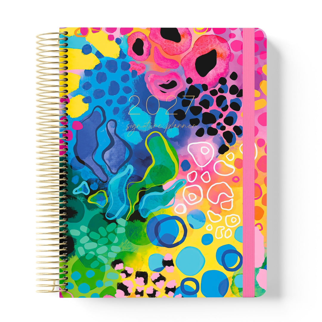 2027 Signature Planner in Boom with bold abstract artwork in pink, yellow, blue and green, gold coil and pink elastic closure.