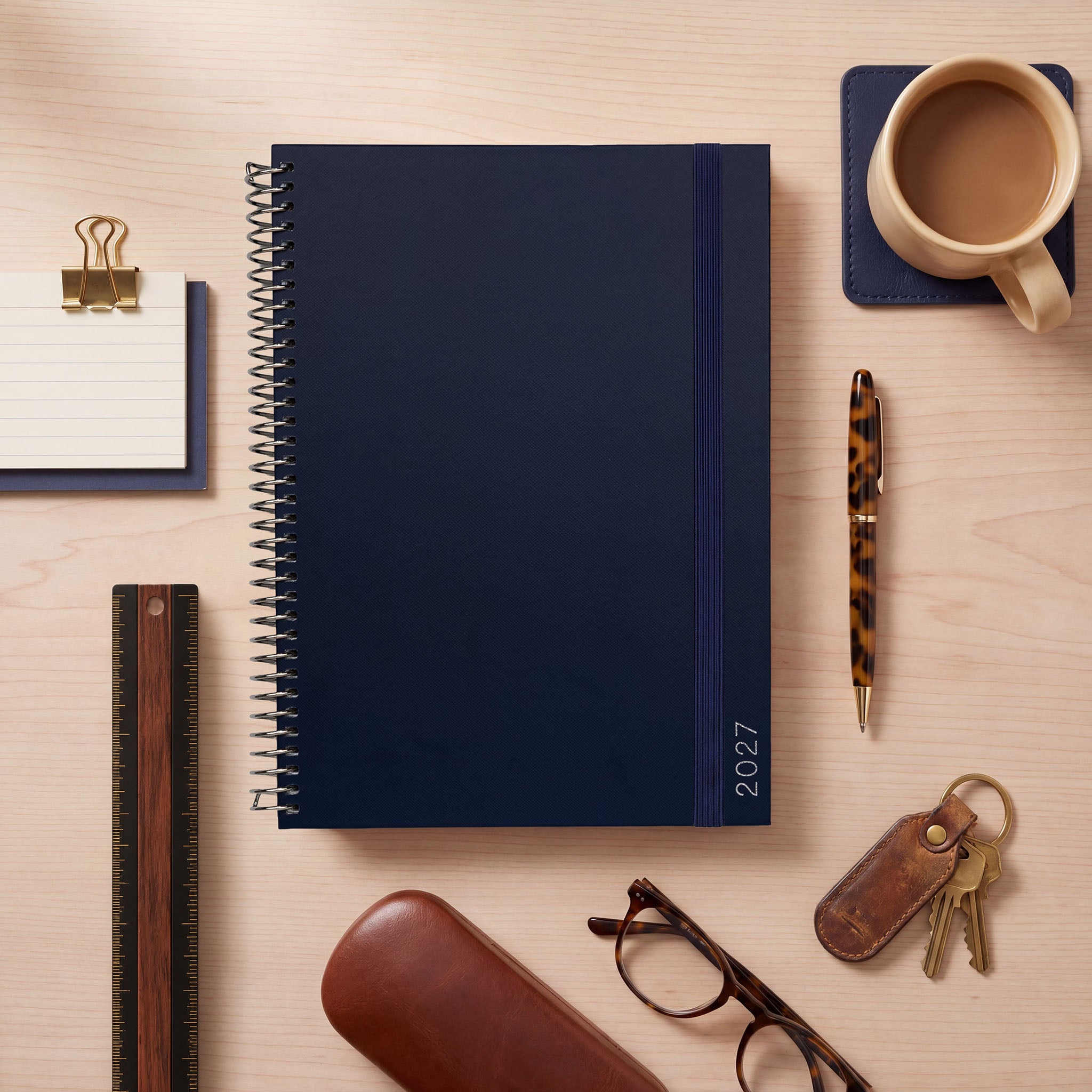 2027 navy slimline planner with elastic closure, styled on a light wood desk with coffee, pen, glasses and keys.