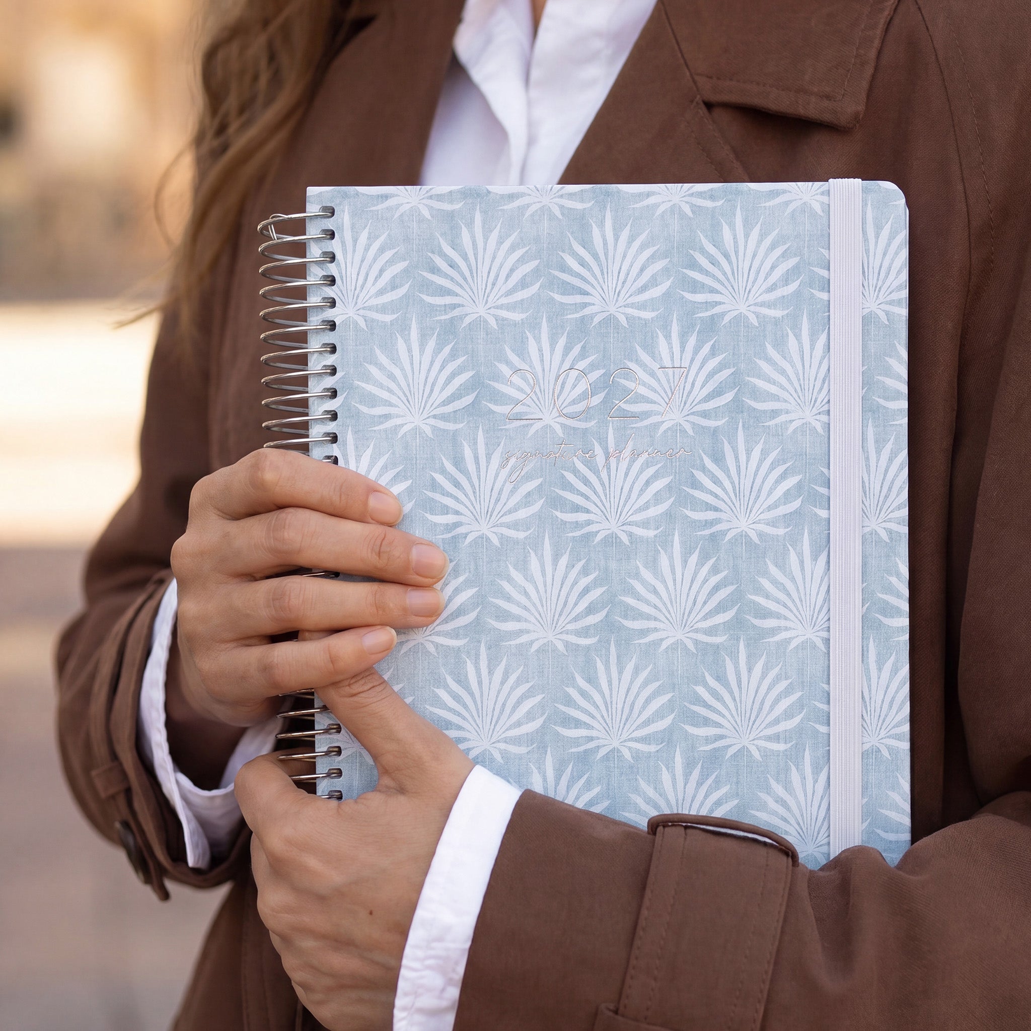 2027 Signature Planner in Palms held by a woman in a brown coat, silver coil and cream elastic closure.
