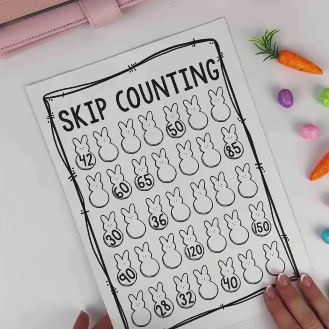 A person holds a completed Easter-themed “DOUBLES” math worksheet featuring brightly colored Easter eggs with solved addition problems inside them. Examples include 8+8=16, 4+4=8, 50+50=100, and 90+90=180. The worksheet has a hand-drawn border and is decorated with pastel colors. Small toy carrots and colorful mini eggs are scattered on the table beside the page.