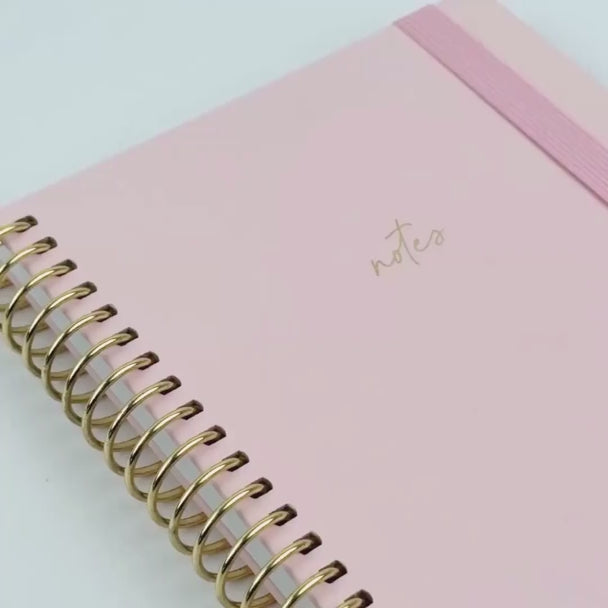 blush notebook with pink elastic and gold binding shown through a close up video