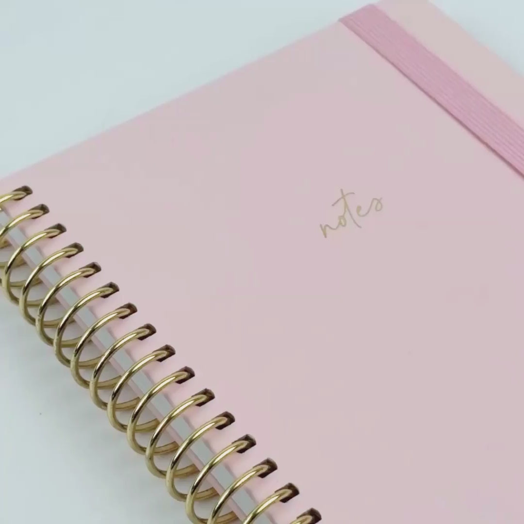 blush notebook with pink elastic and gold binding shown through a close up video