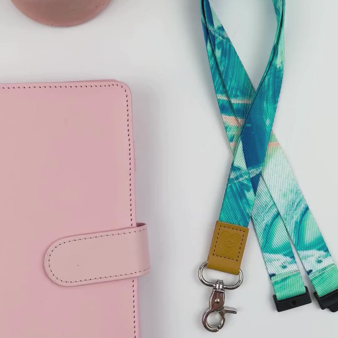 Hands displaying the Paradise island lanyard with soft blue patterned design, silver clasp, and tan leather accent detail.