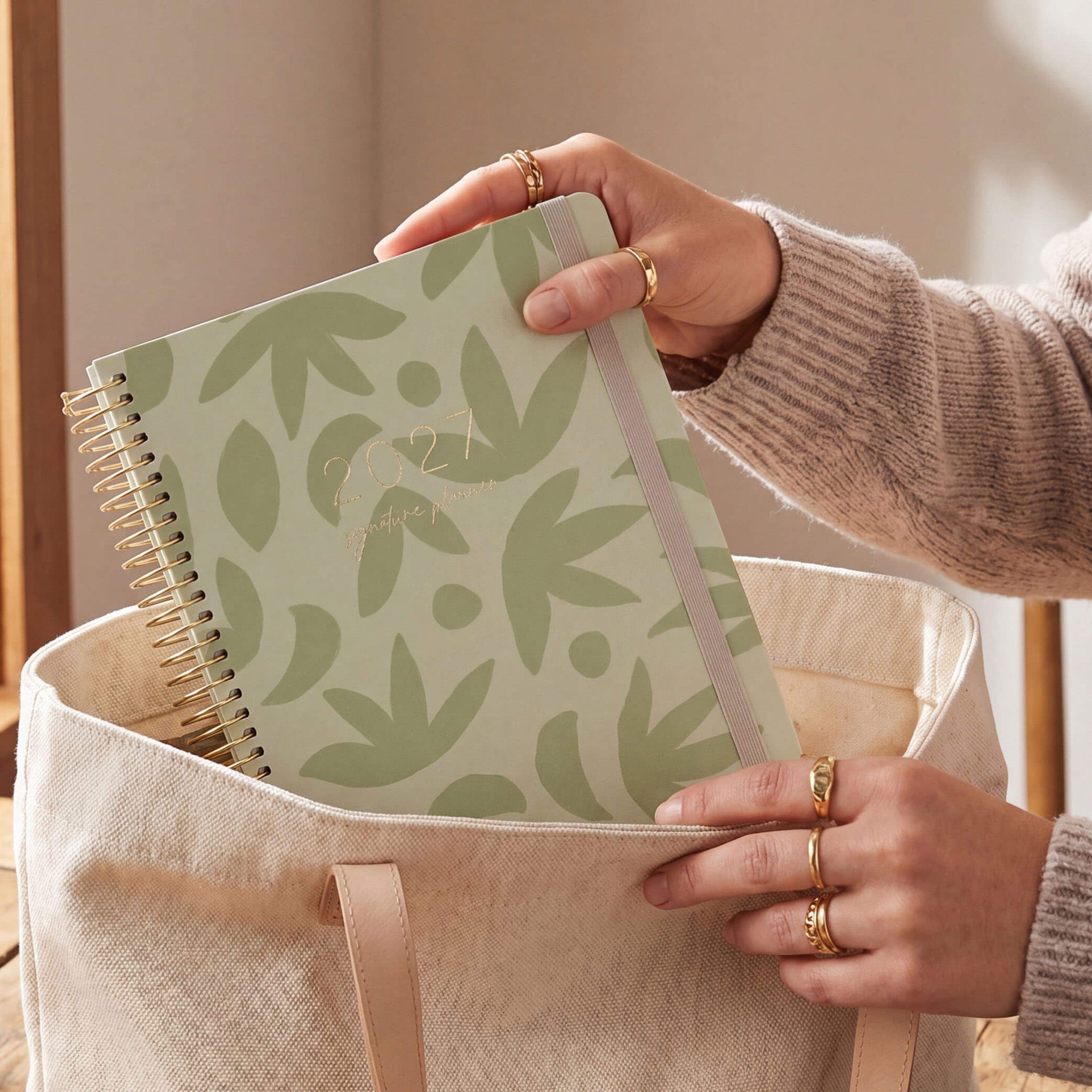 2027 Signature Planner in Sunburst Sage being placed into a canvas tote bag, gold coil and cream elastic closure.