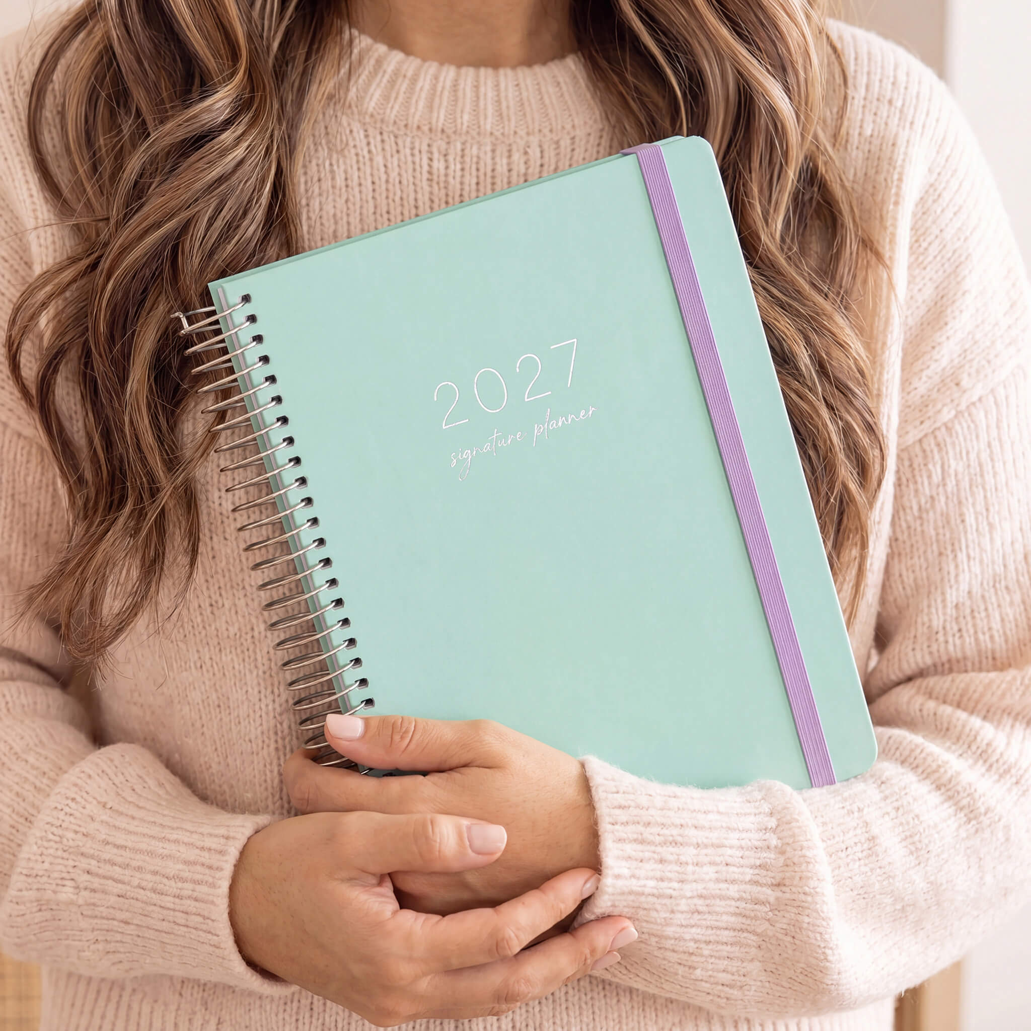 2027 Signature Planner in Seafoam held by a woman in a cream knit sweater, silver coil and lilac elastic closure.
